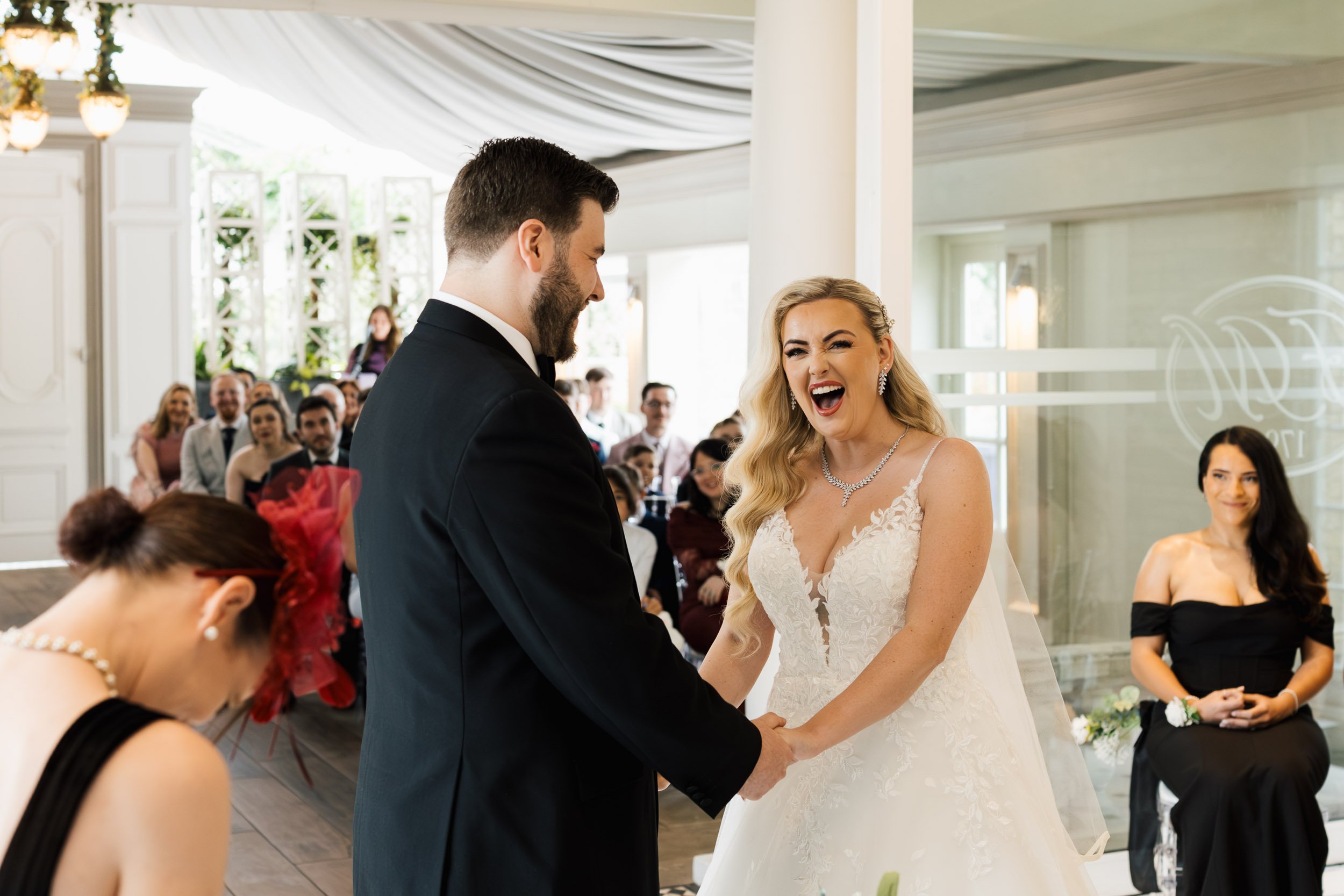 A bride and groom exchange vows during their wedding ceremony inside a decorated event space, with guests seated and watching.