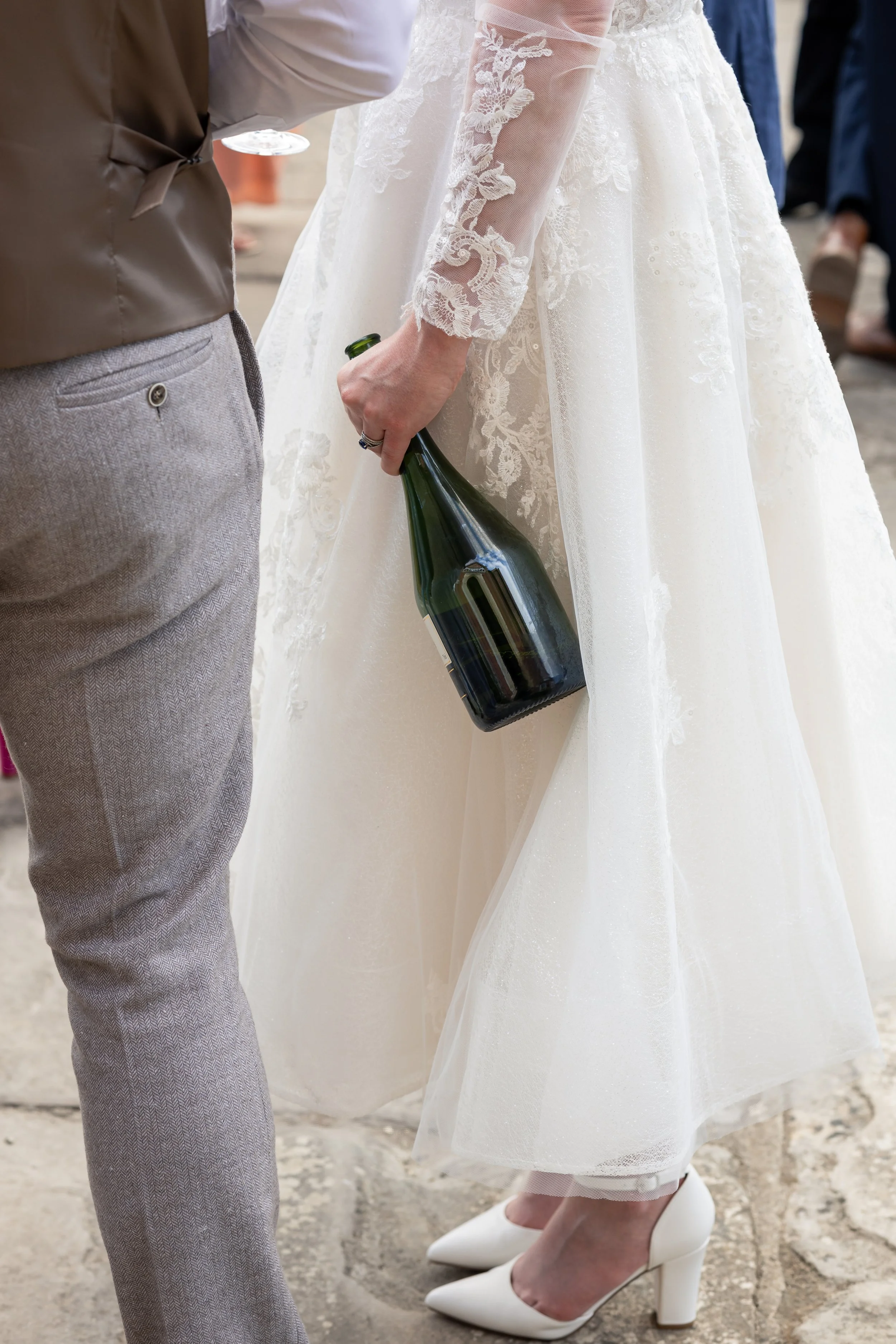 A bride in a lace wedding dress holding a bottle of champagne, standing next to a person in gray pants and a brown vest.