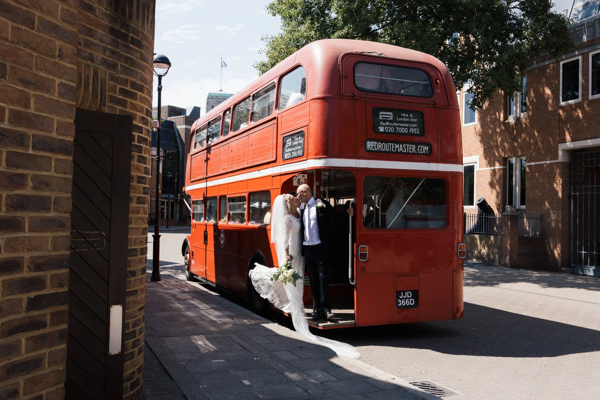 A bride and groom standing together in front of a red double-decker bus during their wedding, with the bride holding a bouquet of flowers and the groom wearing a tuxedo. The scene is set outdoors in an urban area with brick buildings and trees.
