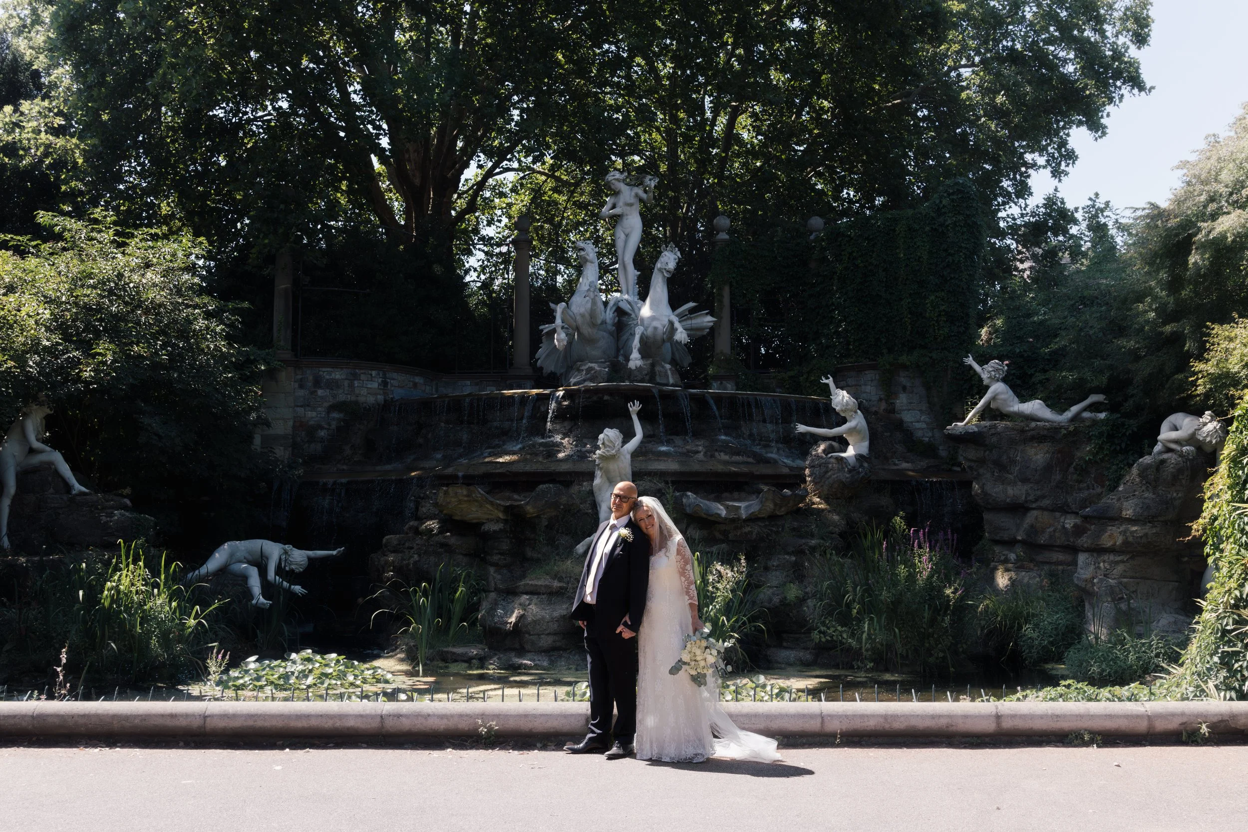 A bride and groom standing together in front of a decorative fountain with statues of mythological figures, surrounded by lush trees and greenery, during a wedding photography session.