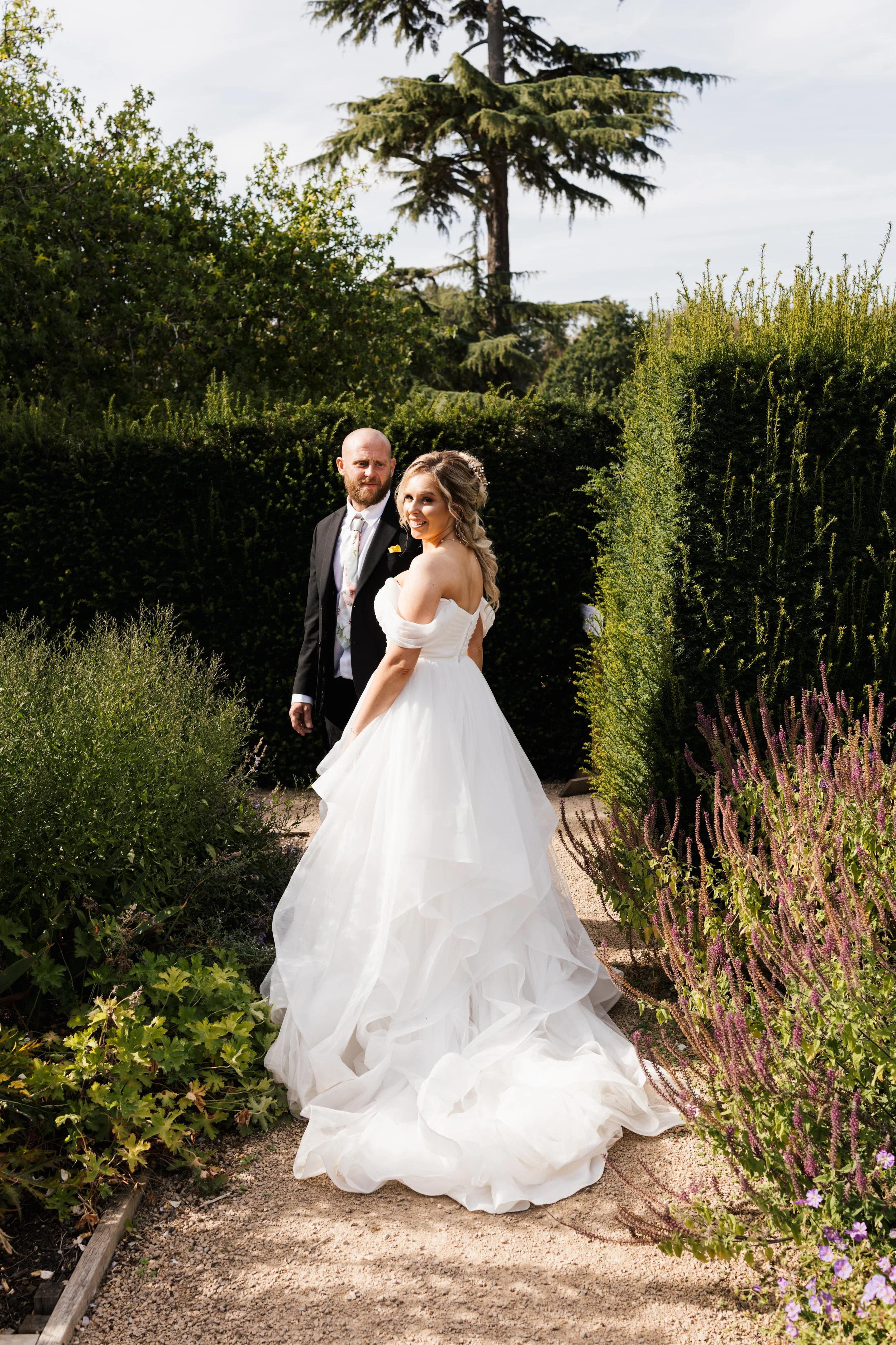 A bride in a white wedding gown with off-shoulder sleeves standing in a garden pathway, smiling, with a groom in a black suit standing beside her, surrounded by lush greenery and purple flowering plants.