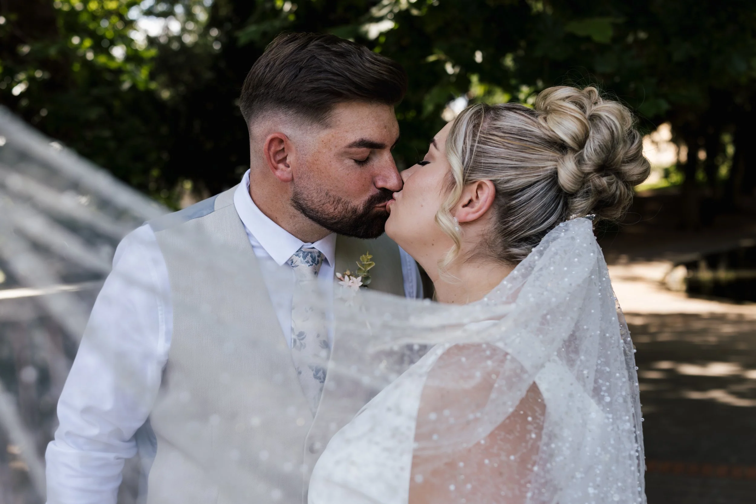 A couple in wedding attire sharing a kiss outdoors, with trees in the background.
