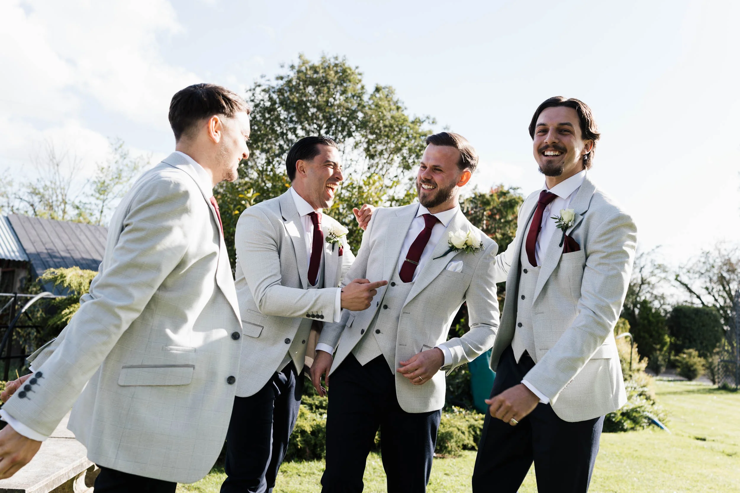 Five men dressed in light-colored suits with dark ties and white shirts, wearing boutonnières, are standing outdoors and smiling at each other during daytime. They are near a tree, with a house and a blue sky with clouds visible in the background.