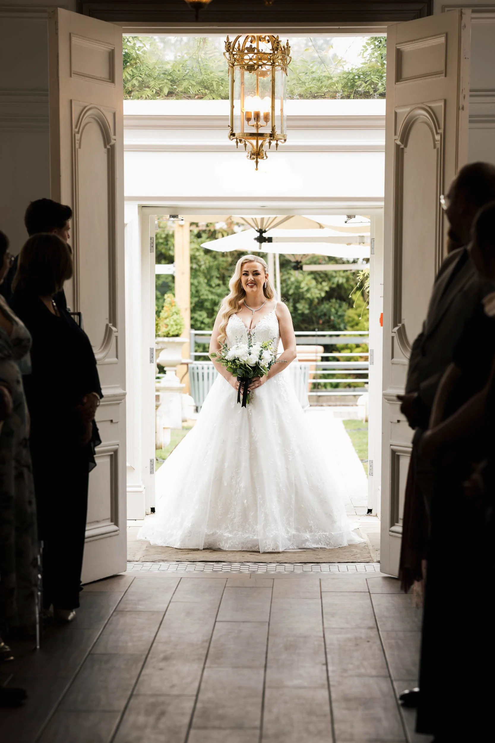 A bride in a white wedding gown holding a bouquet of white flowers standing outdoors, viewed through an open door, with wedding guests in silhouette on each side of the doorway and a chandelier hanging from the ceiling inside.
