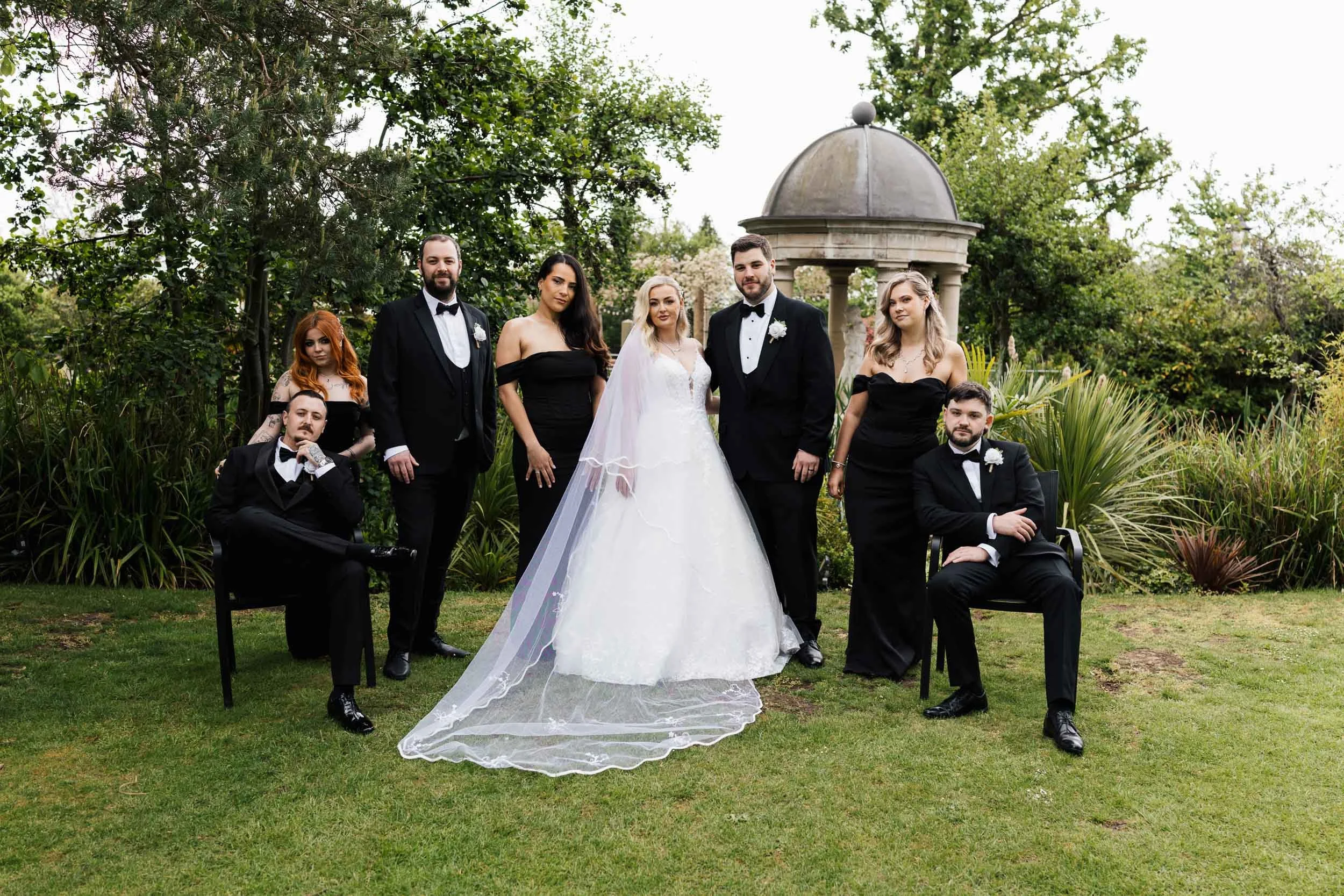 A bride in a white wedding dress with a long veil standing with six well-dressed men and women in black formal attire, outdoors in a garden with greenery and a small stone gazebo.