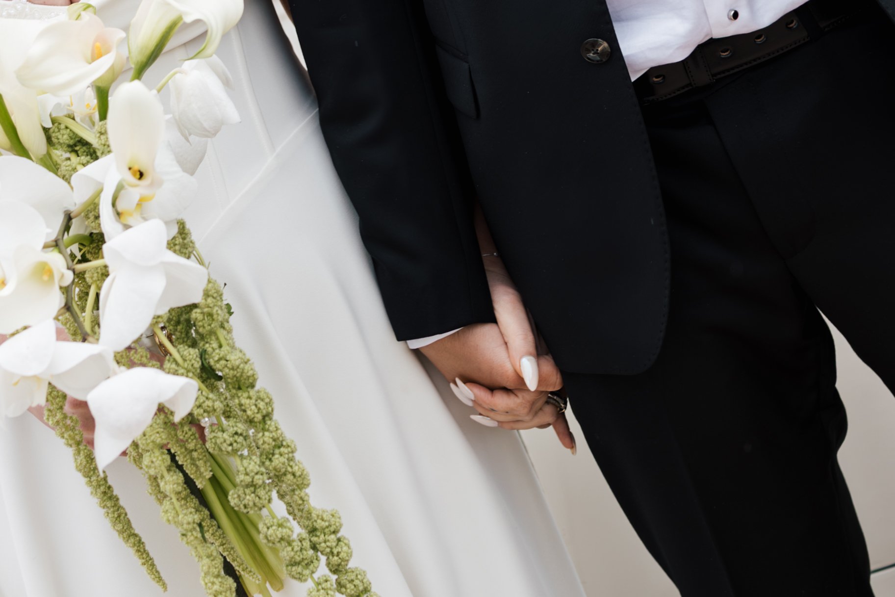 Close-up of a person in a black suit, white shirt, and black belt, with hands clasped in front, sitting at a table decorated with white orchids and green floral arrangements.