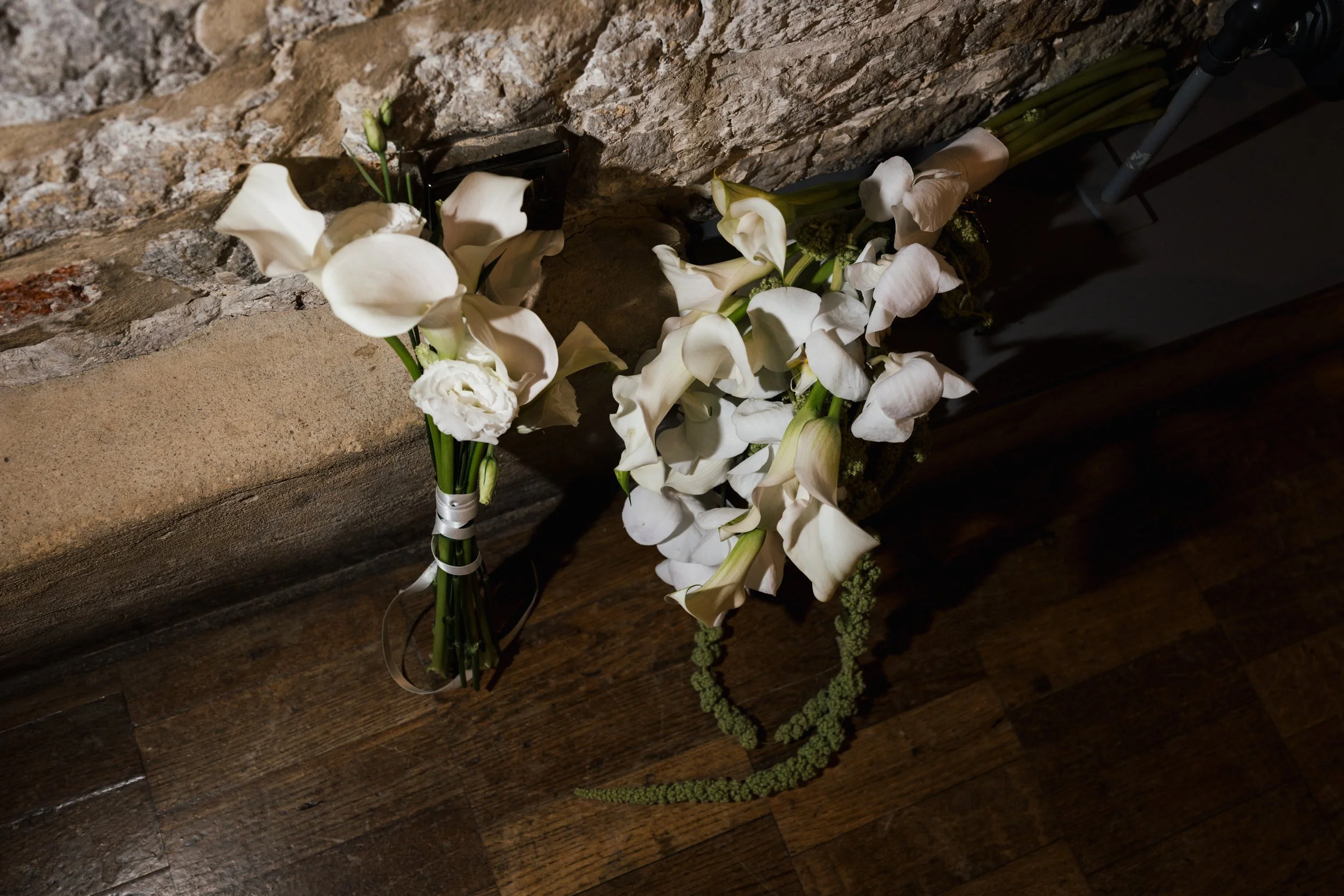 White floral arrangements with calla lilies and other flowers on a wooden surface against a stone wall