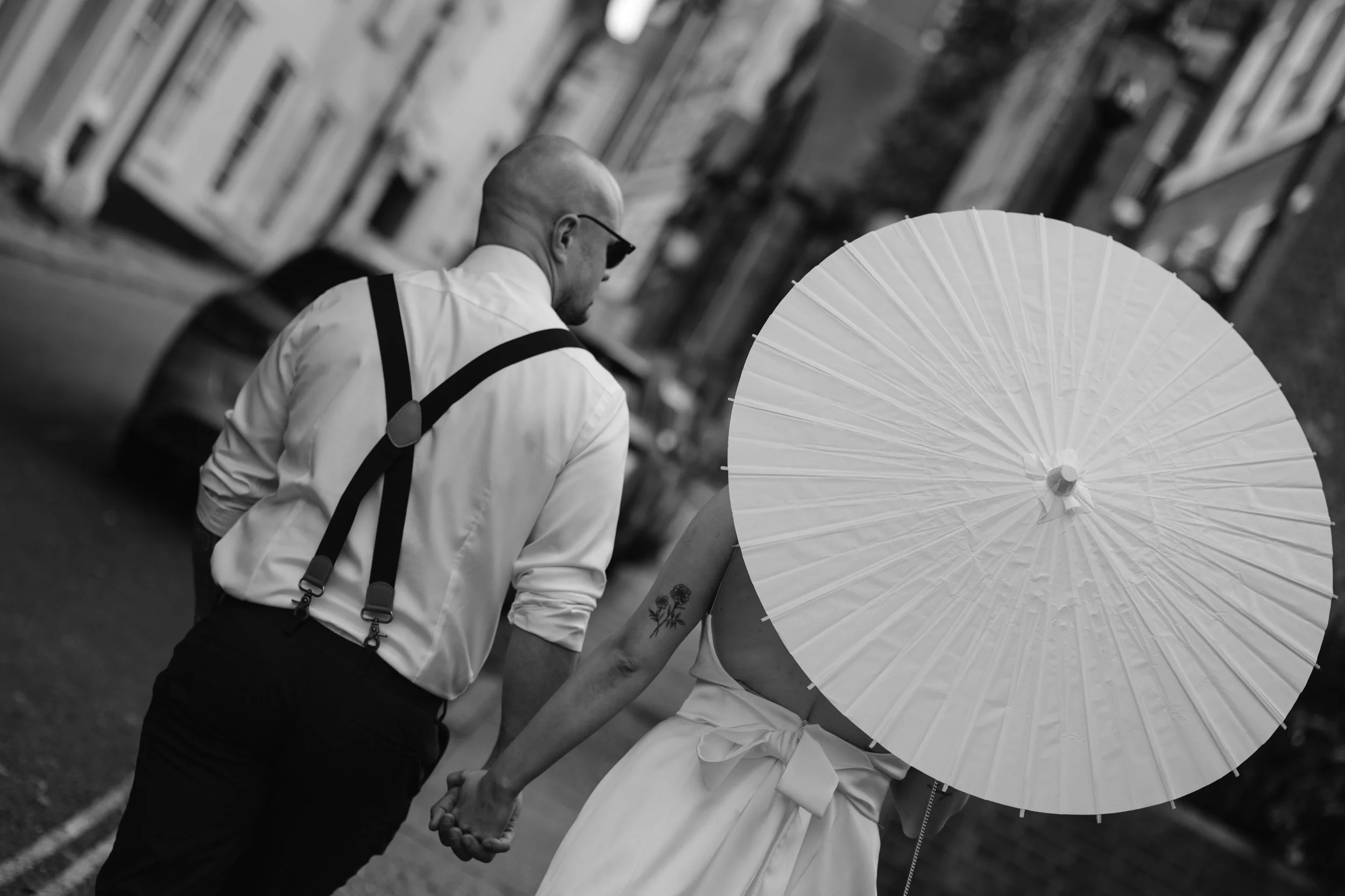 A black and white photo of a couple walking hand-in-hand on a city street. The woman is holding a large white parasol that covers her face and upper body. The man is dressed in a white shirt with suspenders and sunglasses, with a shaved head. They ar
