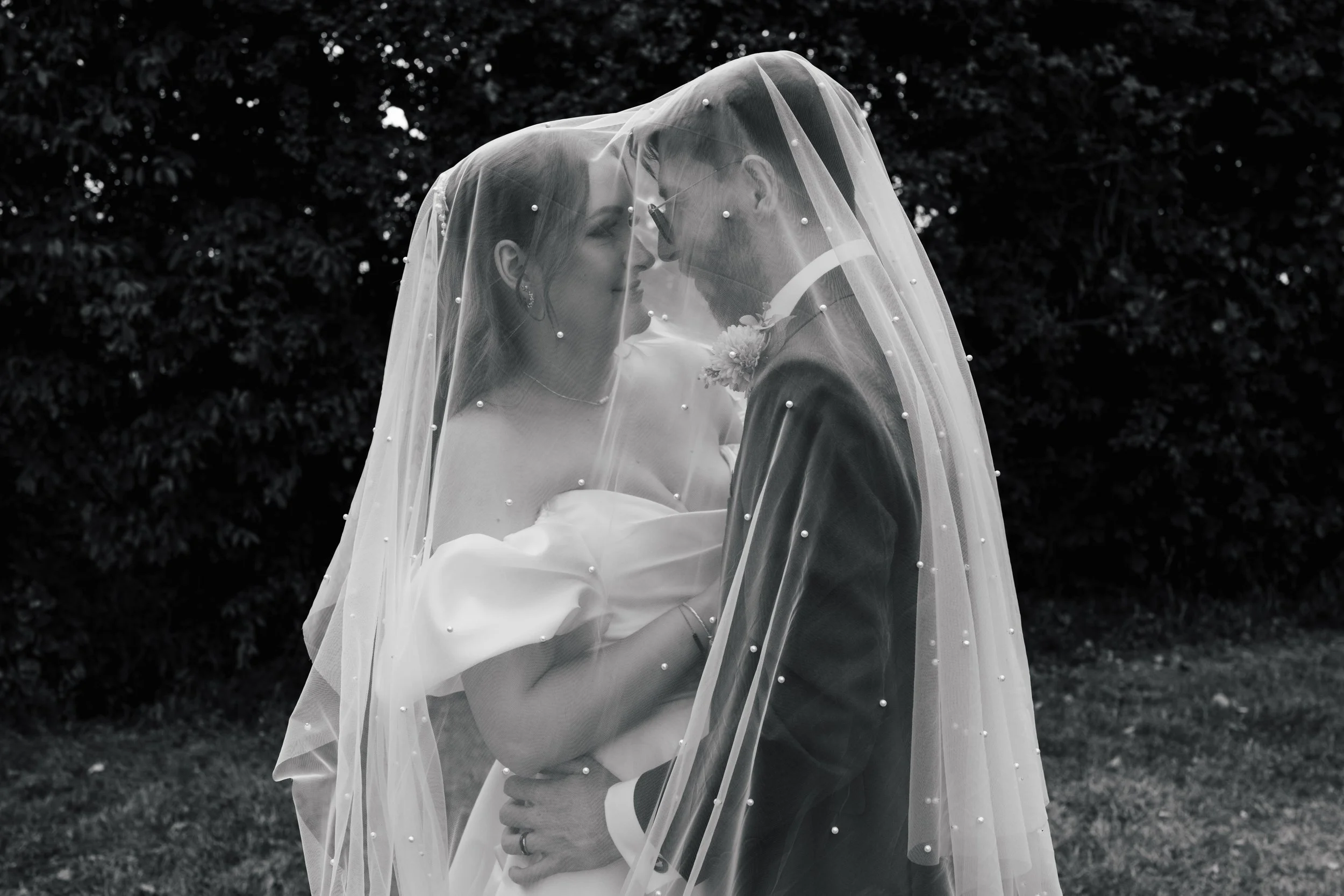 A bride and groom wearing glasses, sharing a close, intimate moment under a veil with pearls, outdoors against a background of dark leaves.