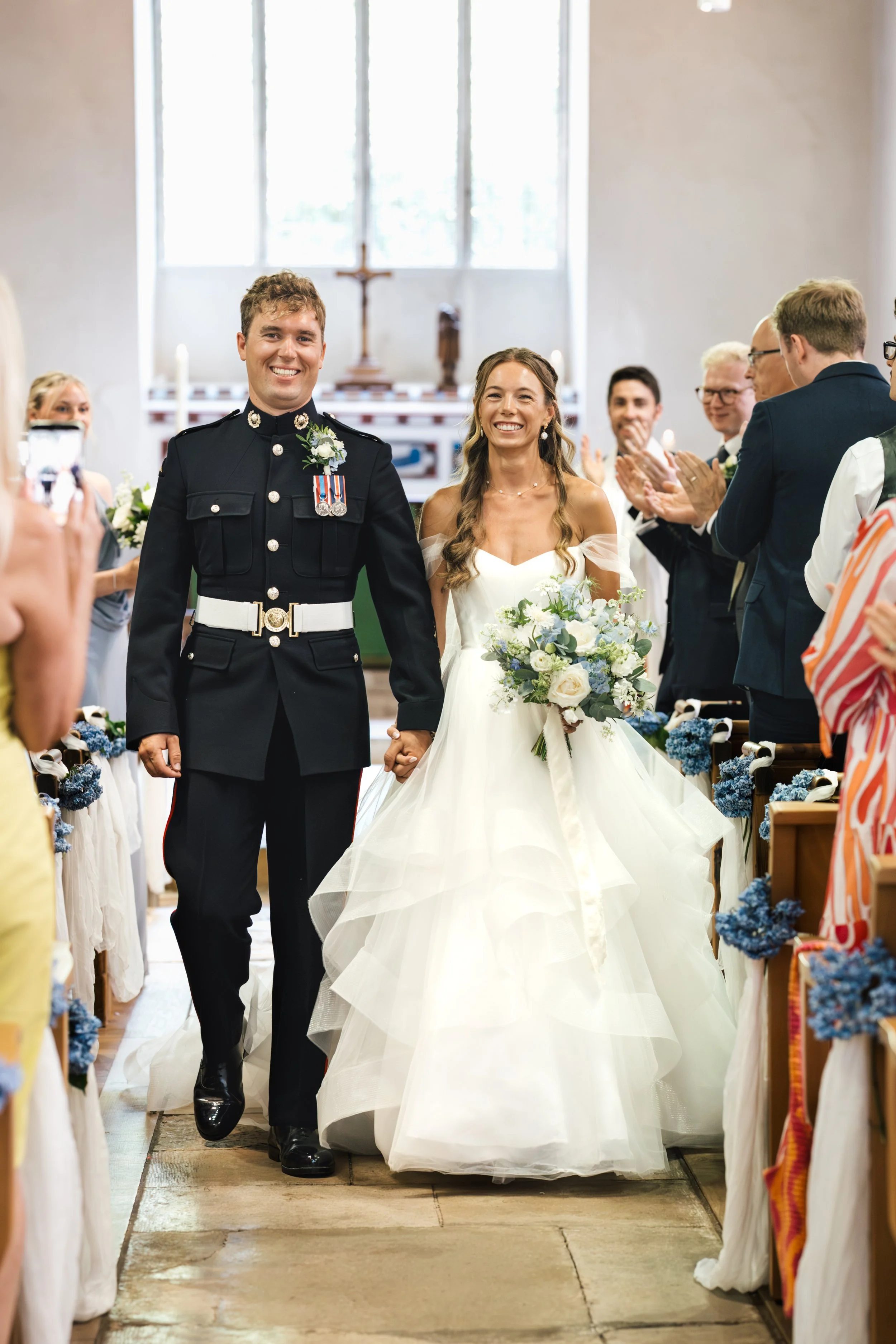 A bride and groom walking down the aisle after their wedding ceremony in a church. The groom is in a military uniform and the bride is in a white wedding dress holding a bouquet of flowers. Guests are clapping and smiling.