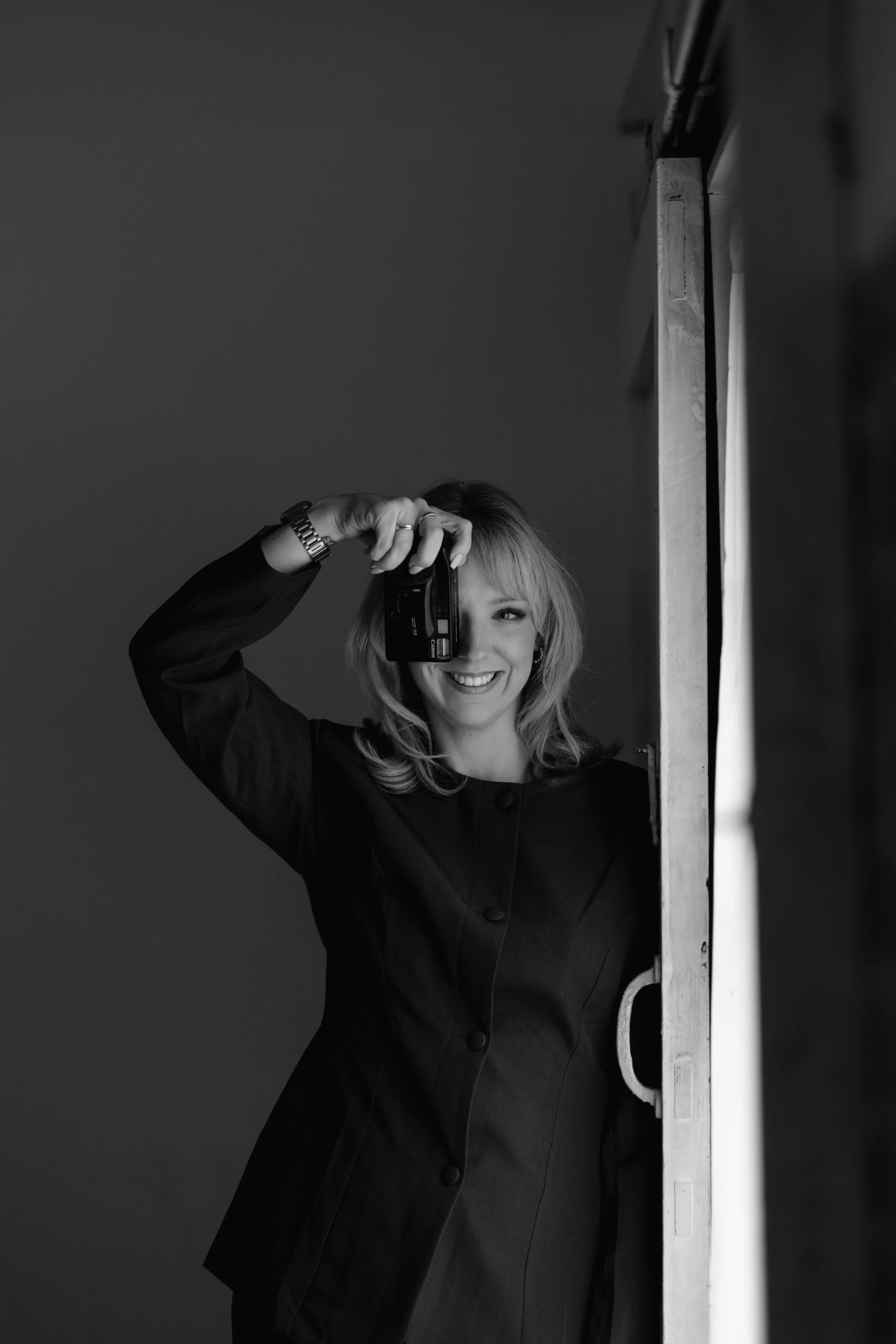 A woman with blonde hair smiling, taking a photograph with a camera. She is standing beside a vertical wooden structure.