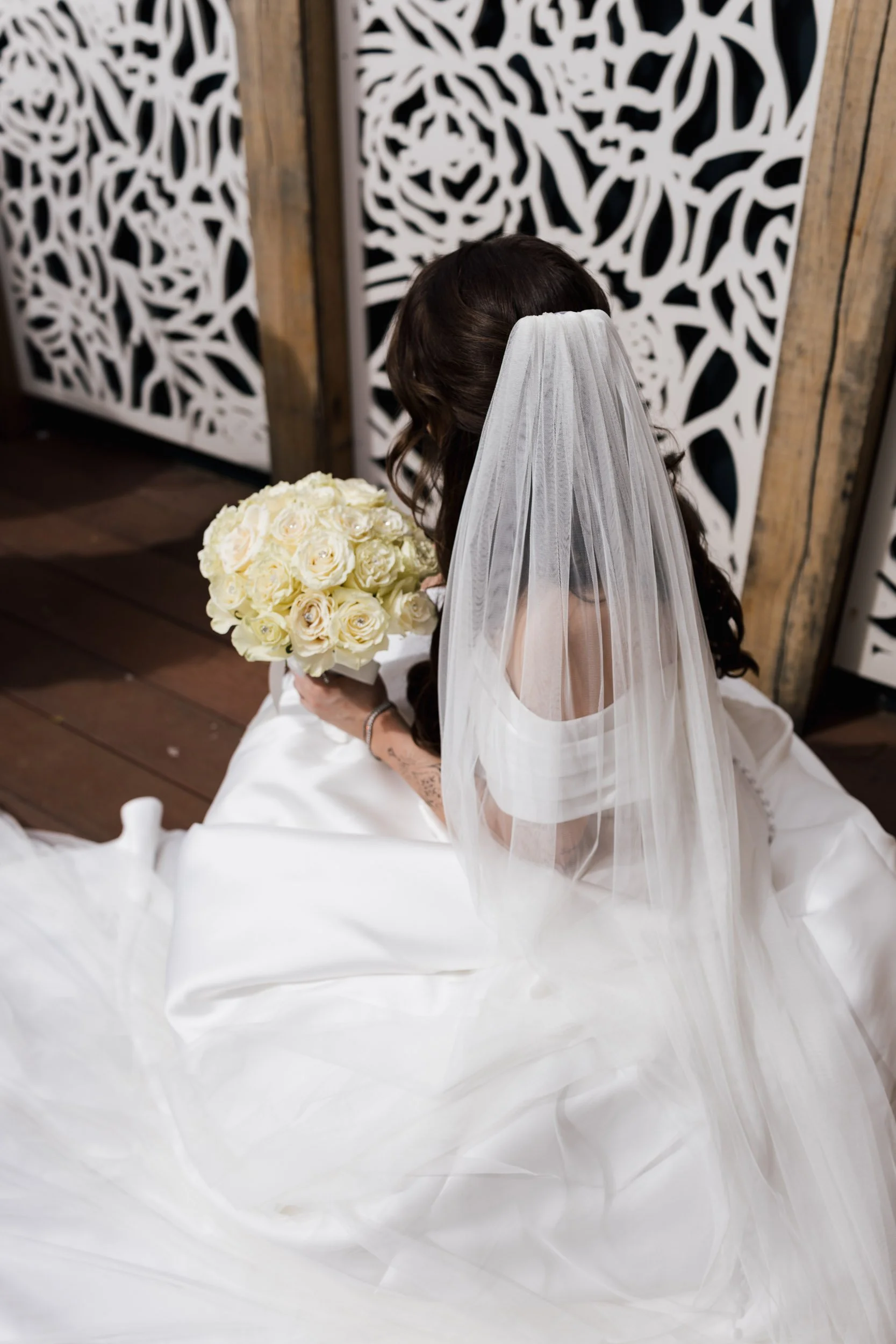 A bride in a white wedding dress and veil, holding a bouquet of cream-colored roses, sitting on the floor with her back turned to the camera, in front of decorative white lattice panels.