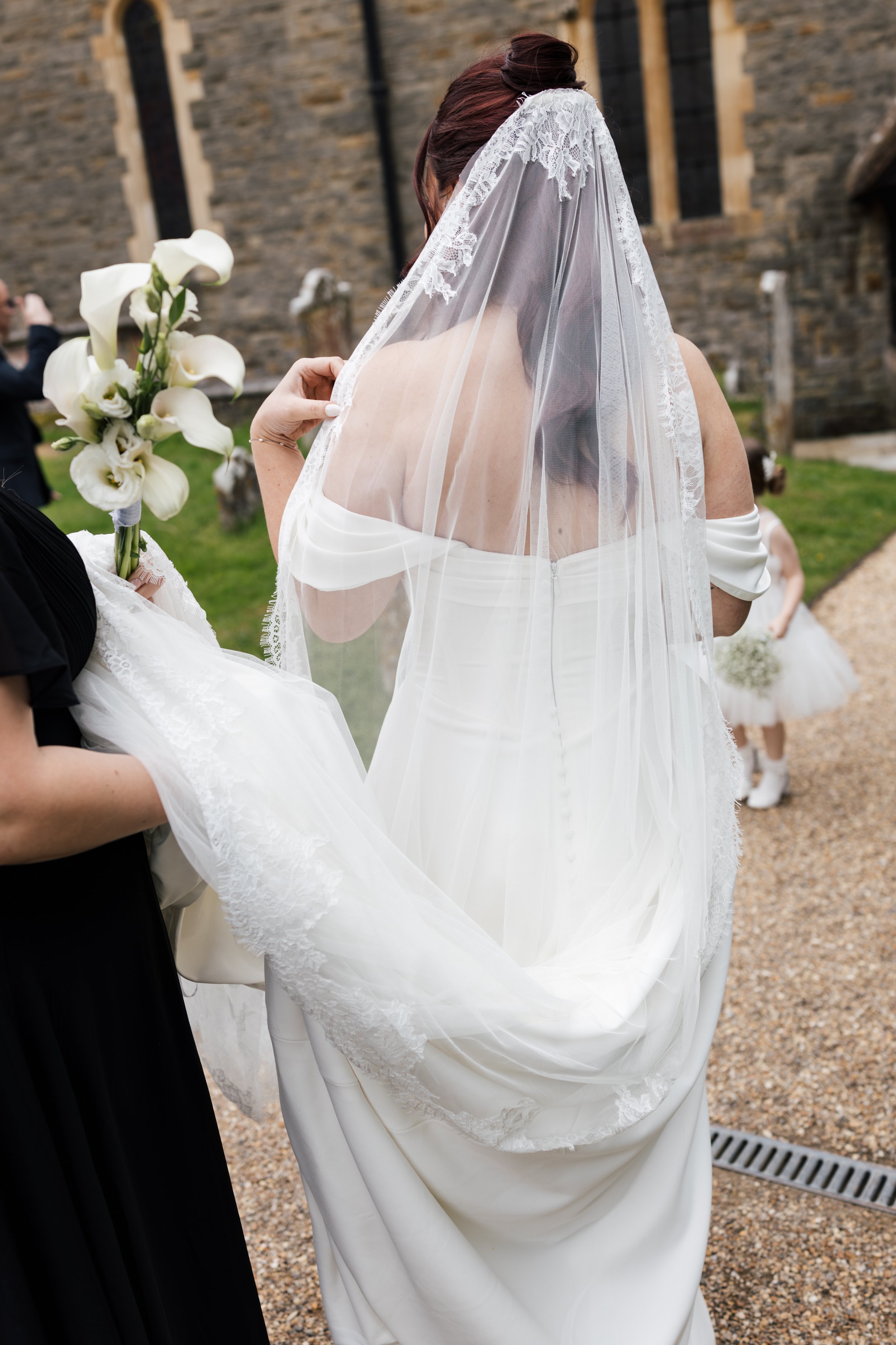 A bride with reddish-brown hair wearing a white off-shoulder wedding dress and a lace veil, holding a bouquet of white calla lilies, standing outdoors near a stone building.
