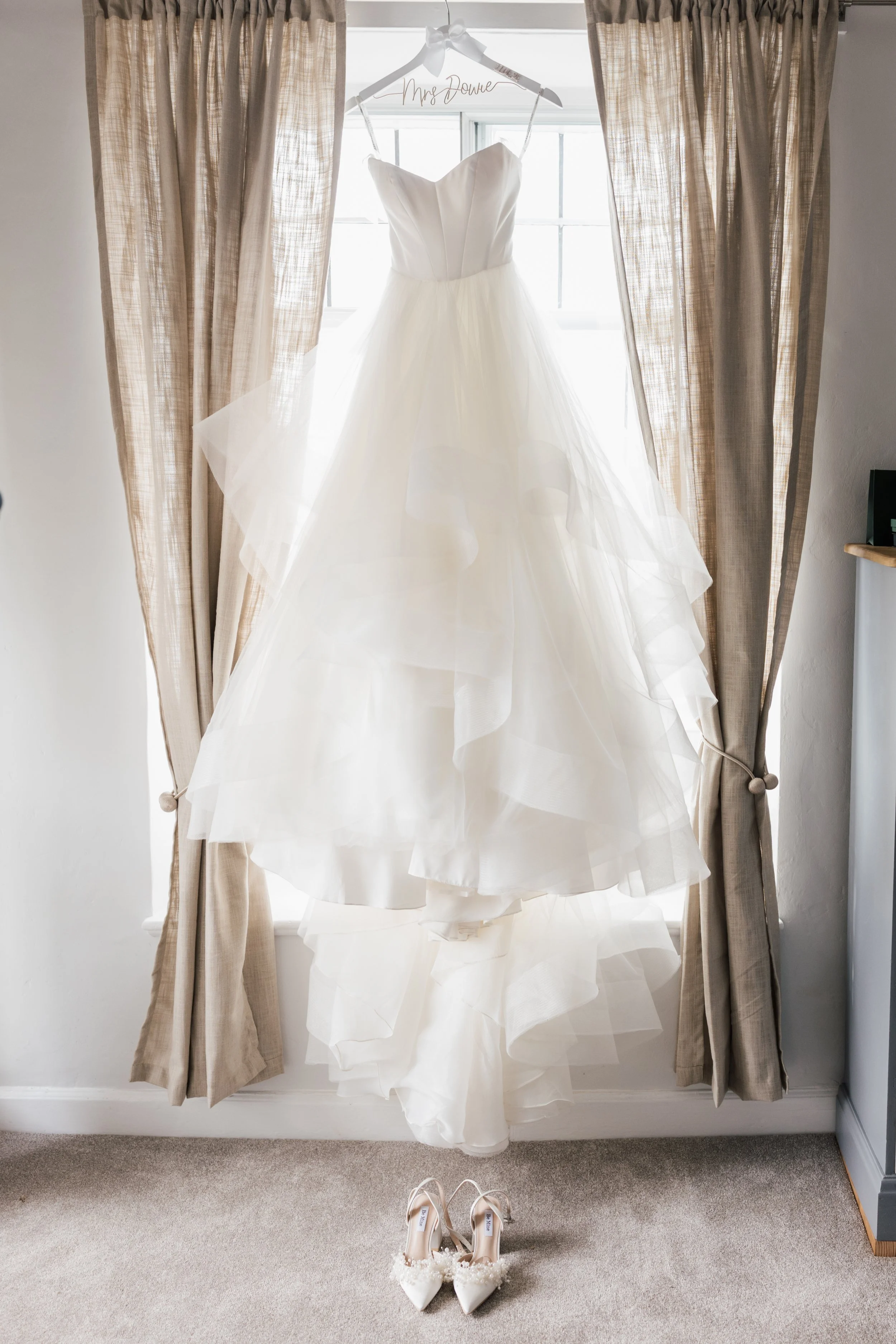 A wedding dress hanging in front of a window with beige curtains, and a pair of white wedding shoes on a beige carpet below.