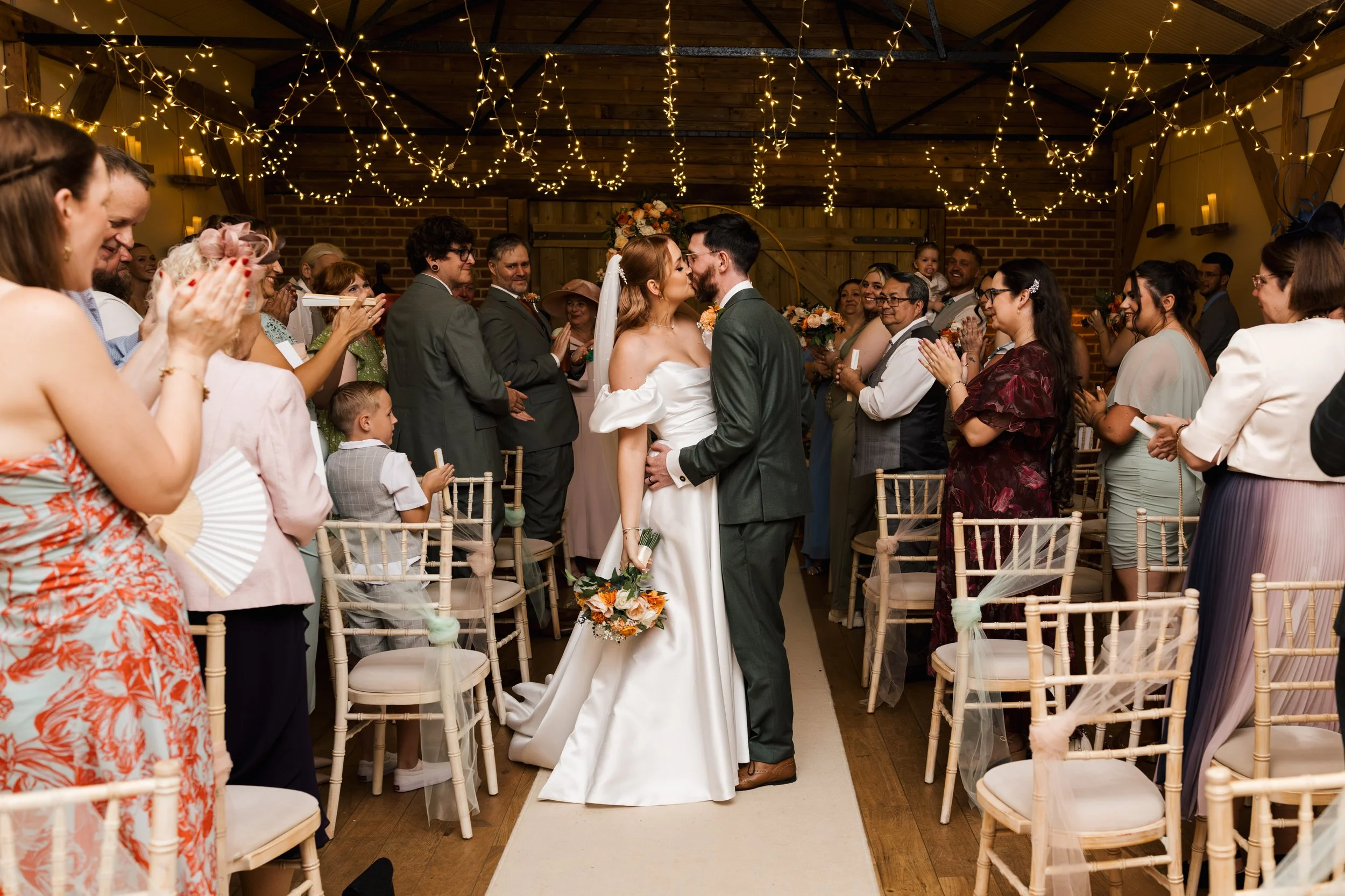 A couple's wedding ceremony with the officiant, bride and groom kissing, guests clapping and smiling, romantic lighting, and rustic wooden decor.