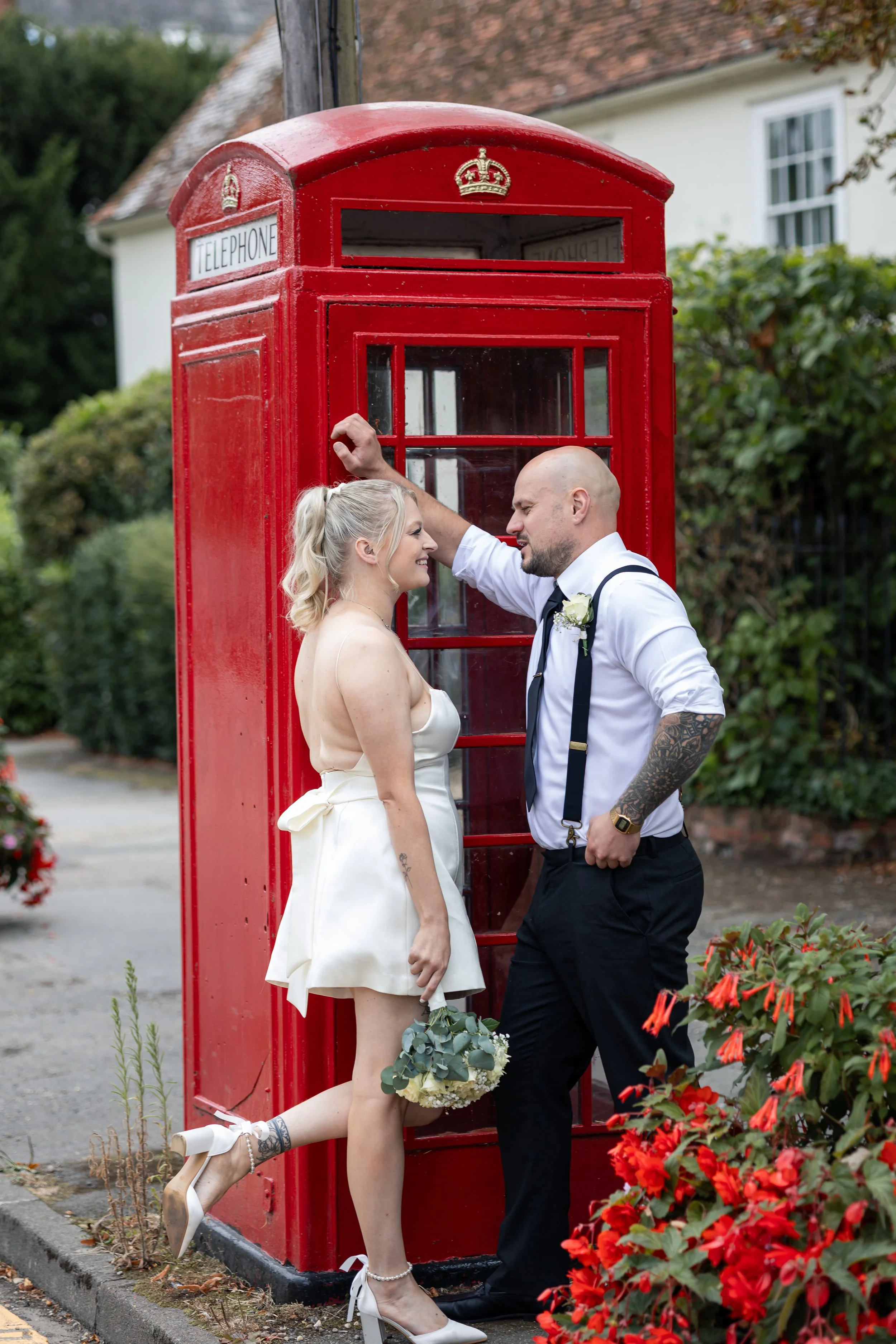 A couple dressed in wedding attire standing next to a red British telephone booth, with the woman holding a bouquet and standing on one foot, as they lean close to each other.