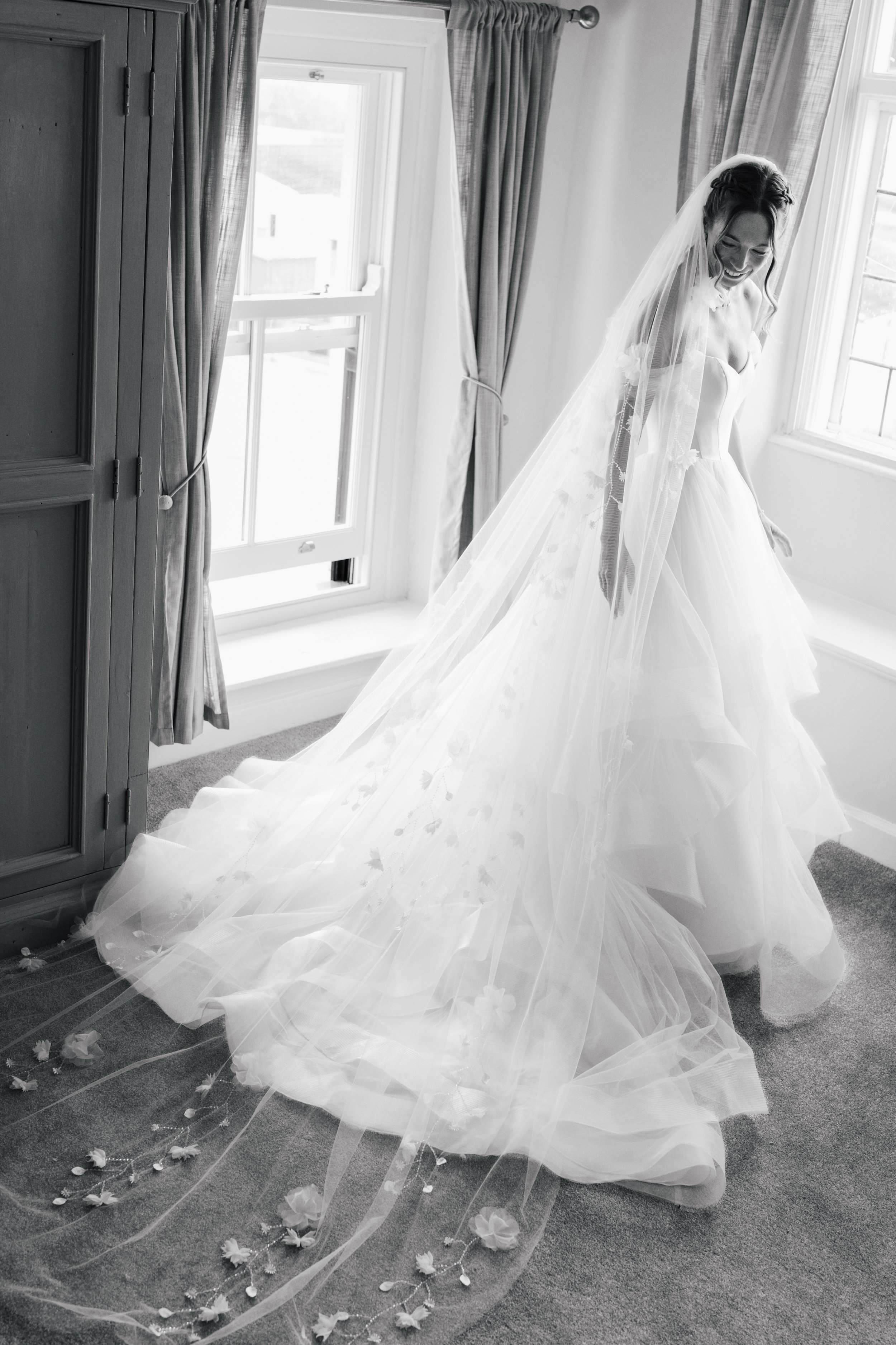 A bride in a wedding dress with a long veil standing inside a bright room near a window, smiling with her eyes closed. The room has curtains and a cabinet, with flower petals arranged on the carpet.