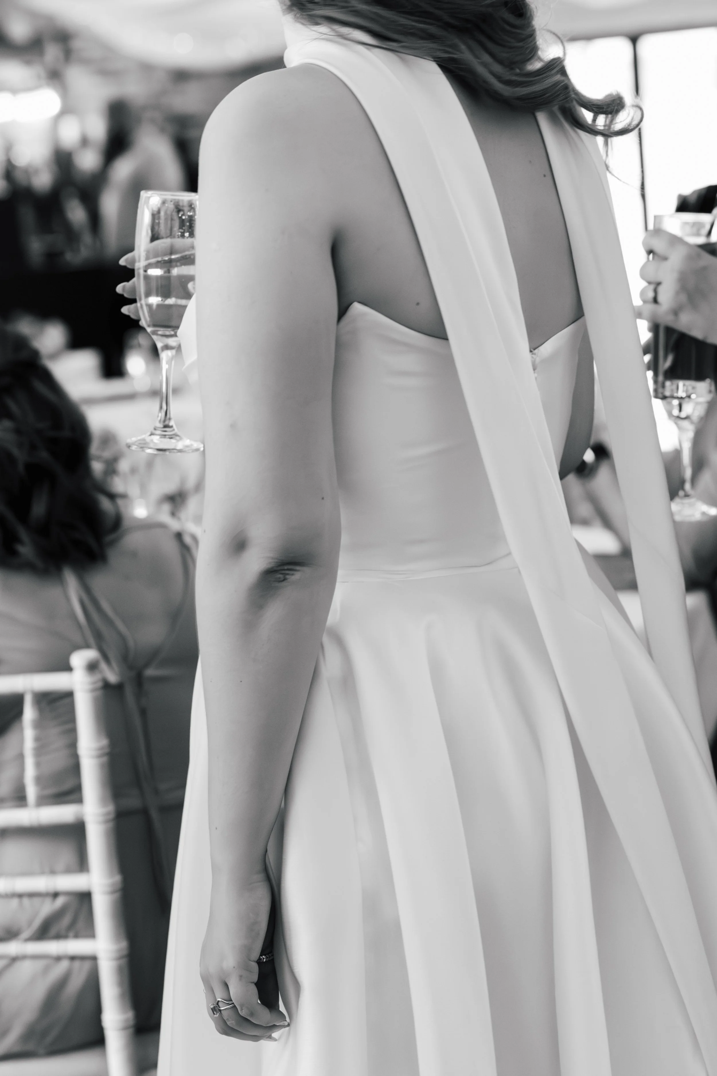 Close-up of a woman wearing a white sleeveless dress, holding a glass of wine, at what appears to be a social or formal event, in black and white.