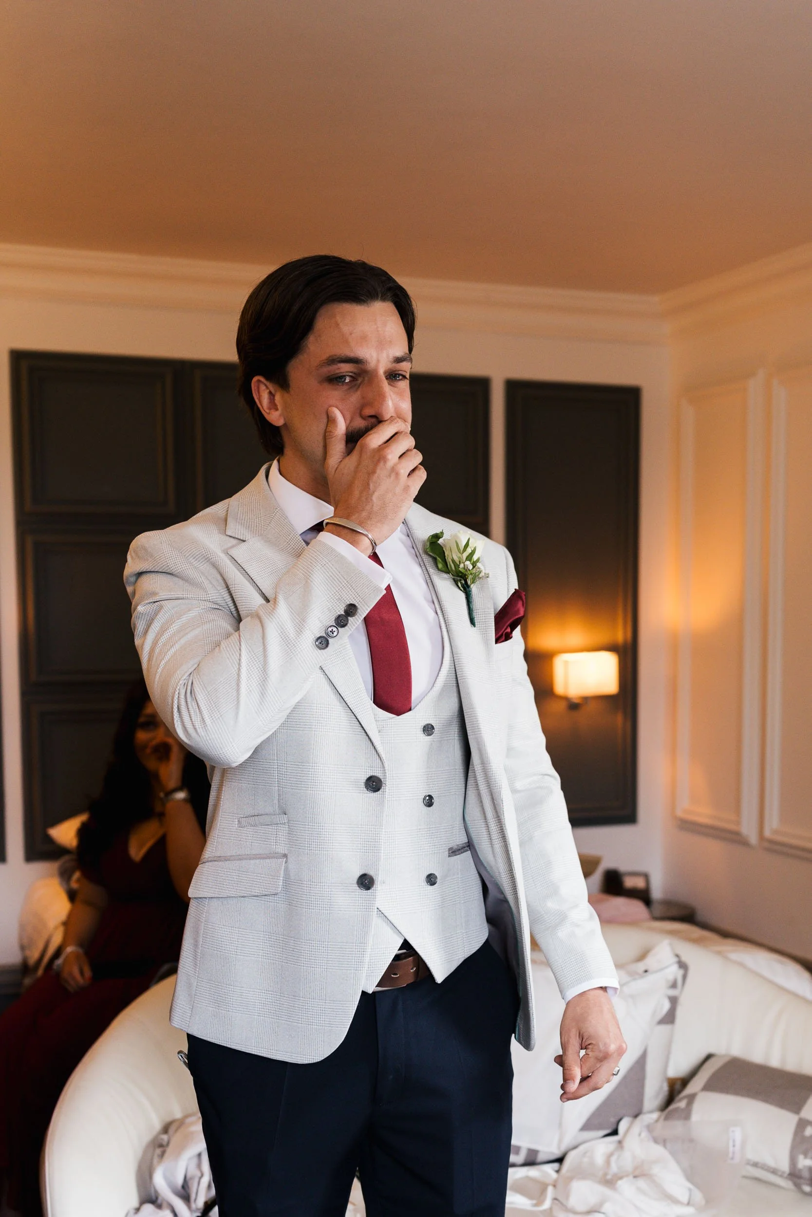 A man with dark hair dressed in a light-colored checkered suit with a white shirt and red tie, holding his face with his hand. Behind him, a woman is sitting on a beige couch, wearing a dark top, in a room with dim lighting and black wall decorations