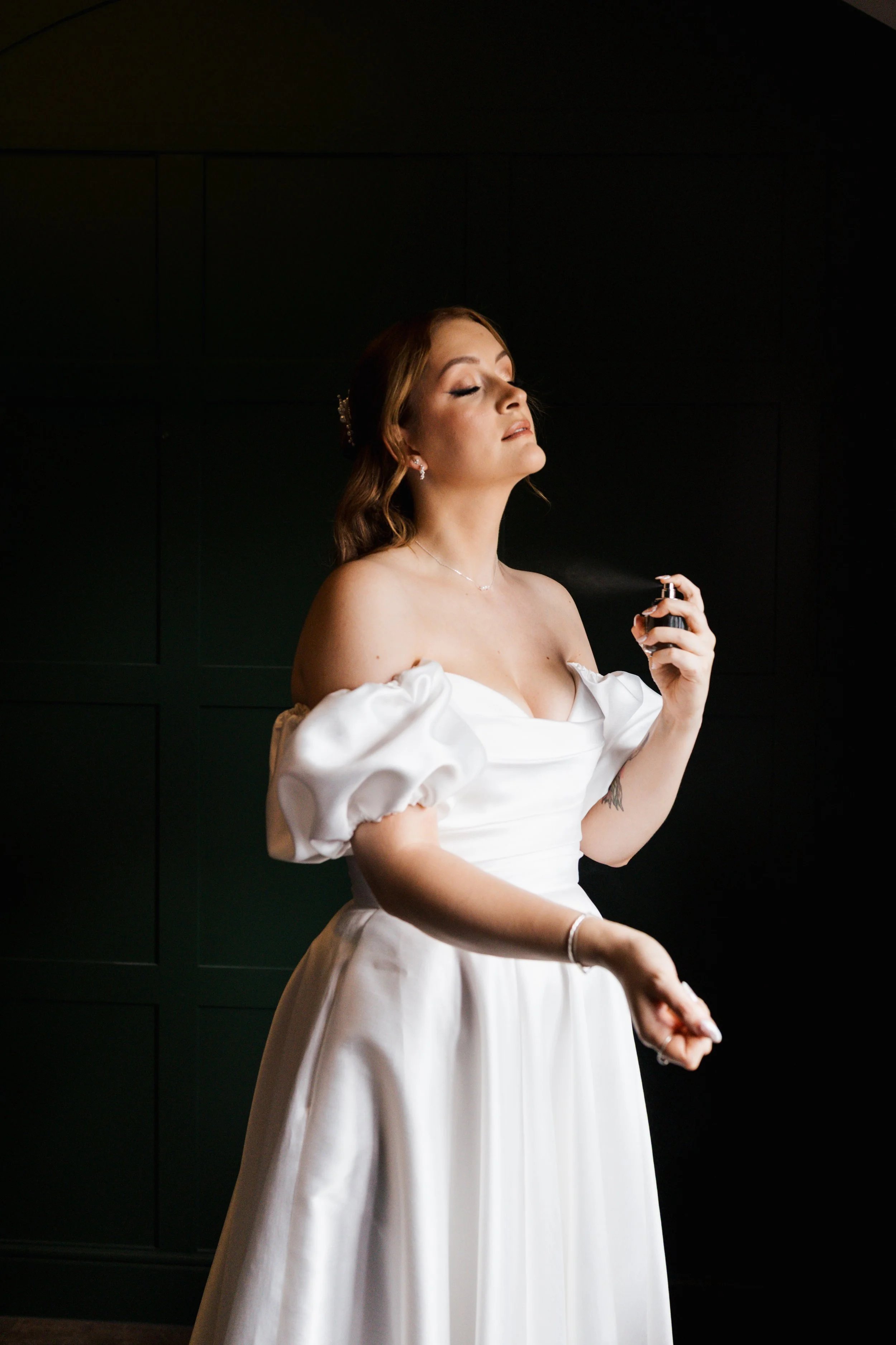 A woman in a white dress with puffy sleeves is spraying perfume on herself, with her eyes closed against a dark background.