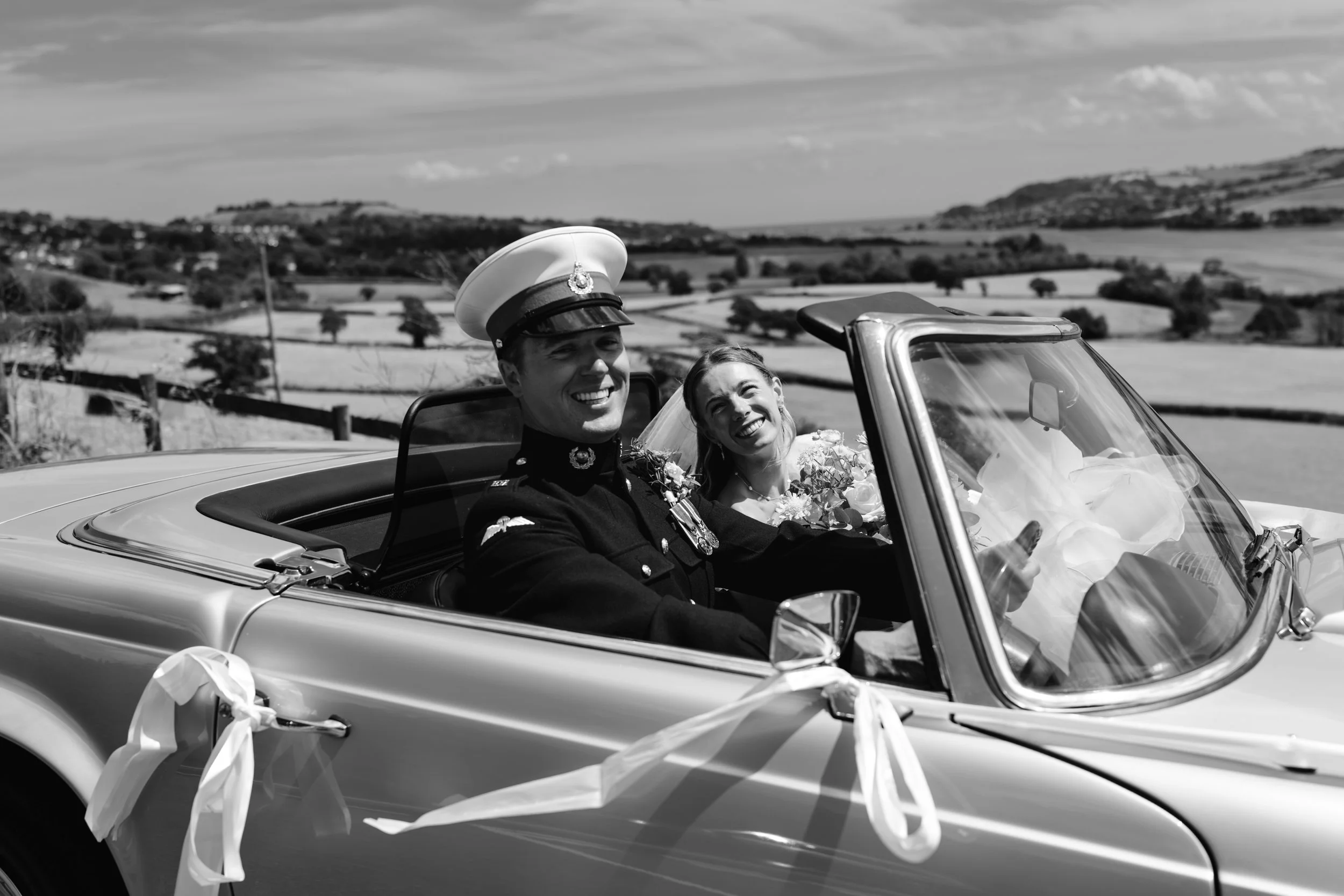 A black and white photo of a smiling bride and groom in a vintage convertible car, with the groom wearing a military uniform and hat, wedding decorations on the car, in a rural countryside setting.
