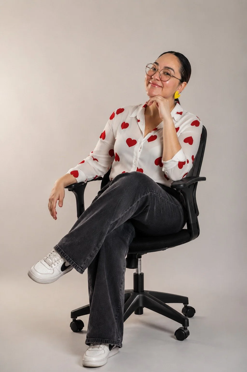 Woman sitting in an office chair, wearing a white shirt with red hearts, black jeans, and white sneakers, smiling and resting her chin on her hand.