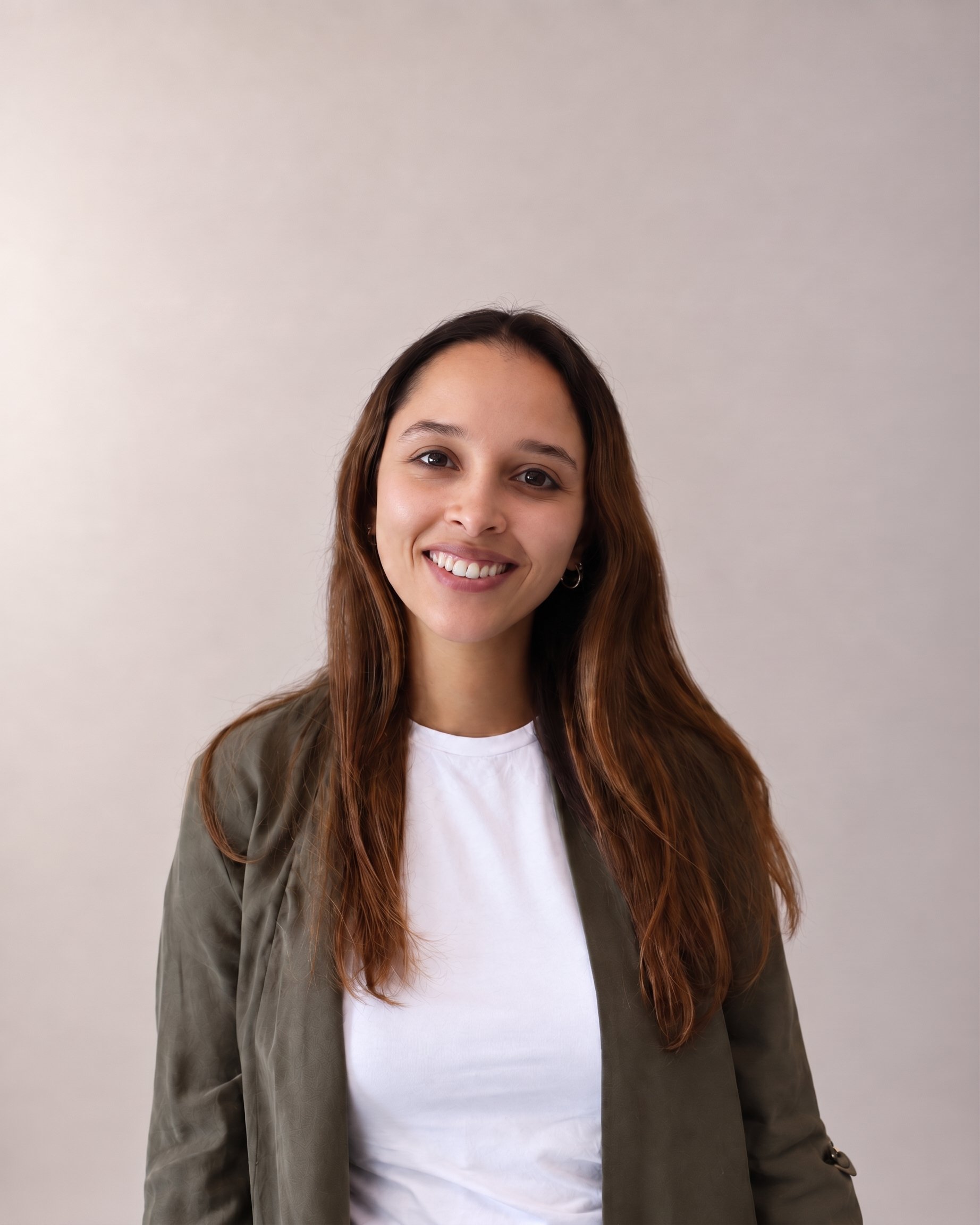 A smiling woman with long, wavy brown hair wearing a white shirt and an olive-green jacket standing against a plain, light-colored background.