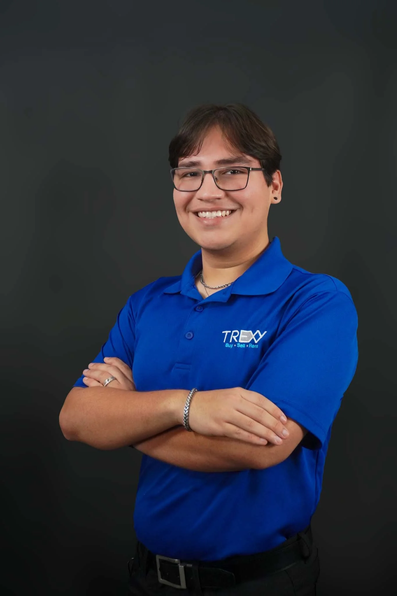 A young person with short dark hair, glasses, and a black earrings with a cross, smiling and crossing arms, wearing a navy blue shirt with small white pattern, standing against a white background.