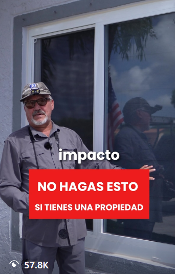 Man in sunglasses and gray jacket standing outside next to a window with a reflection of an American flag.