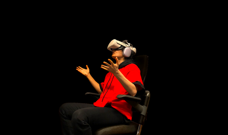 A person in a red shirt sits in a chair wearing a VR headset and headphones, gesturing astonishment with both hands raised, against a black background.