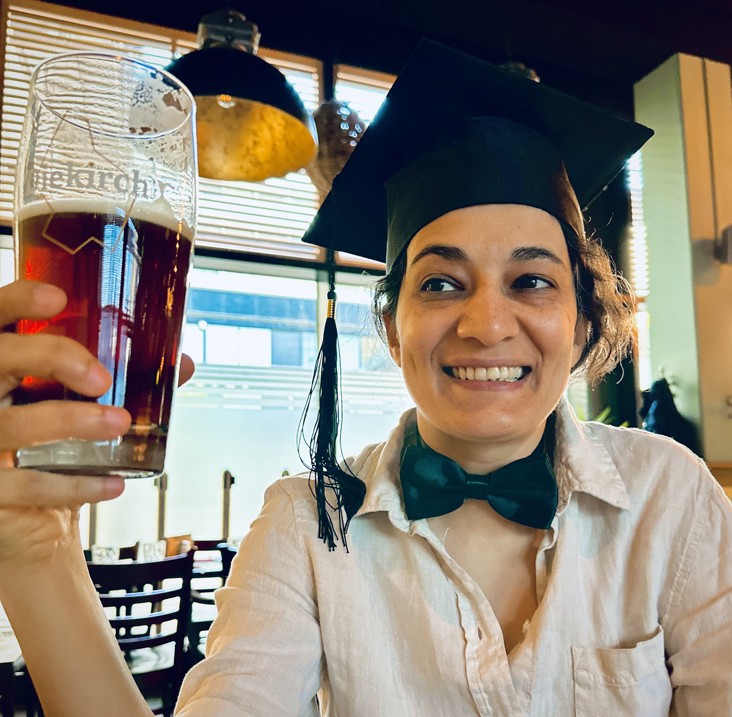 A smiling Sahar Niknam wearing a black graduation cap and a dark green bow tie raises a large glass of beer in a toast, seated inside a restaurant.