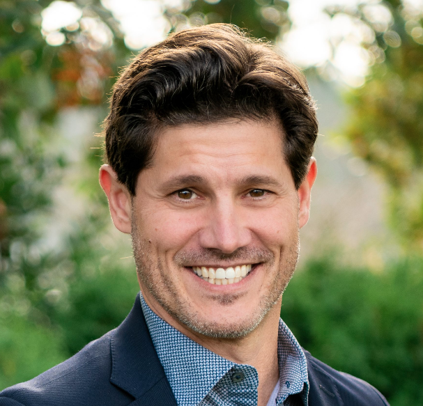 A man with dark hair and a beard smiling outdoors, wearing a blue shirt and dark blazer, with greenery and blurred orange flowers in the background.