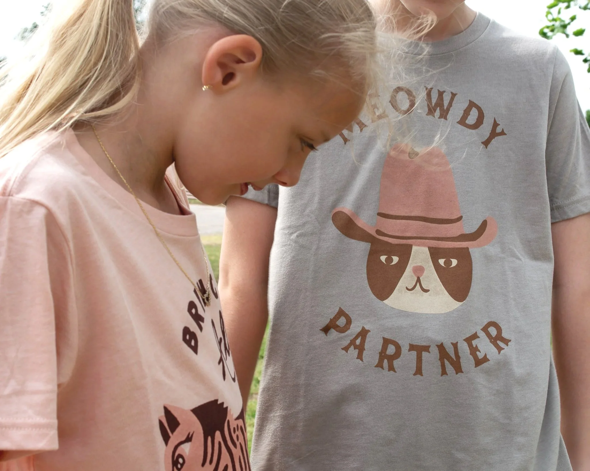 Close-up of two children standing outdoors, looking down, with trees in the background. One girl has blonde hair, wears a light pink shirt with black lettering and a gold necklace. The other child, partially visible, wears a gray shirt with a graphic of a dog and the word "friends."