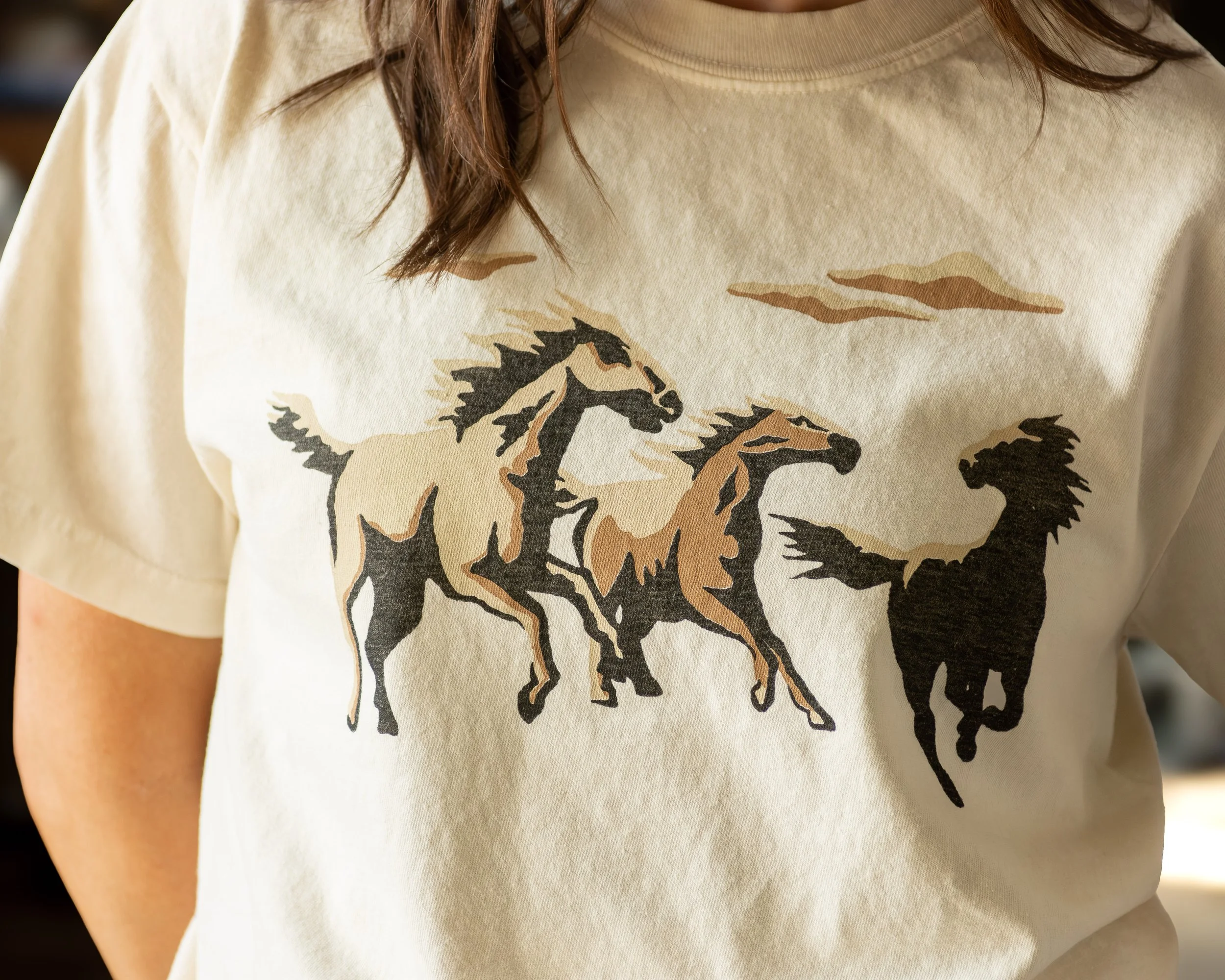 Close-up of a person wearing a beige T-shirt with a printed design of two brown and black horses running.
