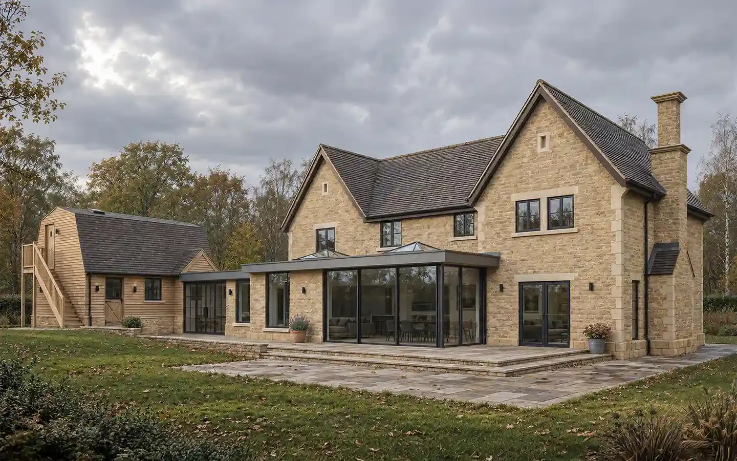 Rear garden view of a Cotswold stone house with a contemporary flat-roofed glazed extension and adjoining timber-clad garage with partial conversion.