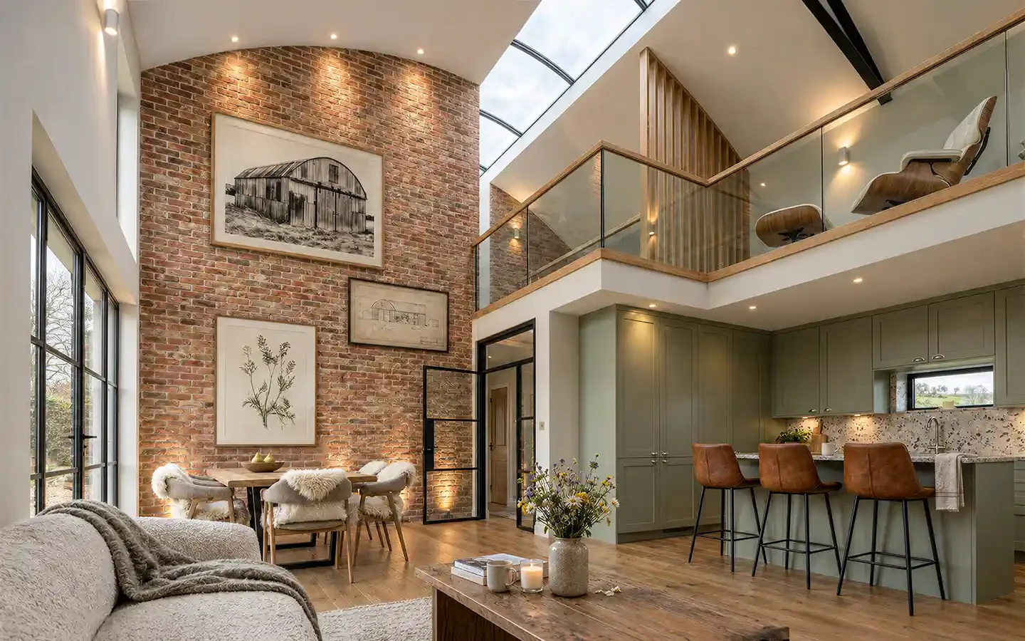 Interior of a Dutch barn conversion in Solihull with an open-plan kitchen, dining and living area, exposed brickwork, double-height ceiling, roof glazing and a mezzanine with glass balustrades.