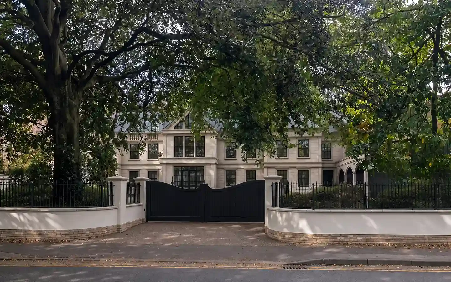 Street view of a bespoke replacement home in a Solihull conservation area, with a pale stone façade, black entrance gates and mature trees behind a boundary wall.