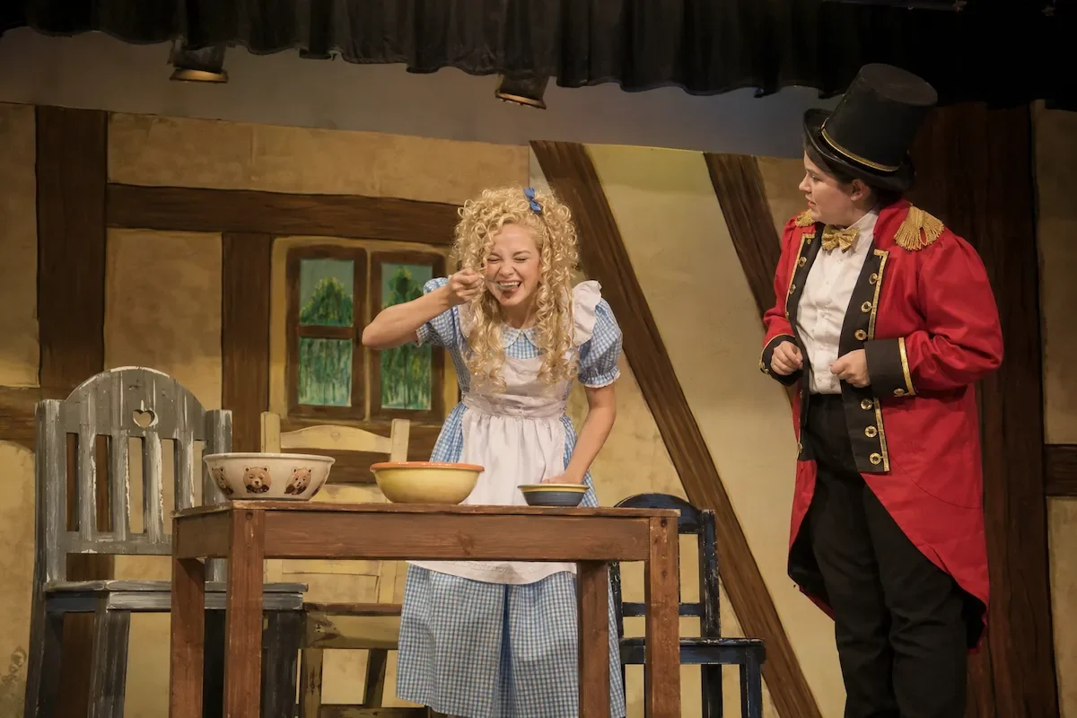 Goldilocks tries some of the porridge at the bears house as her friend Ringo looks on