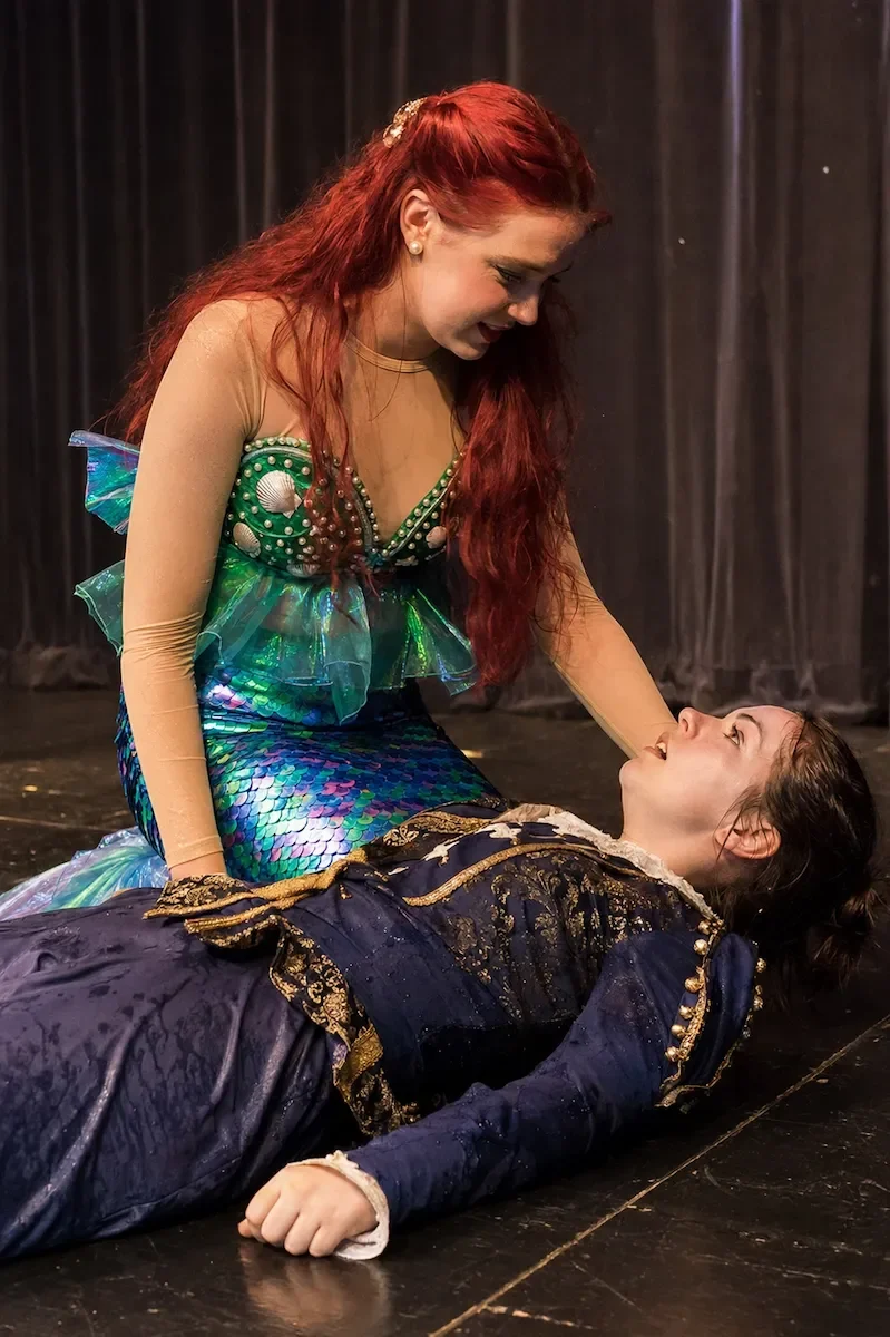 Two women dressed in costumes, one, the Prince, lying on the floor and the other, as Ariel, leaning over her, holding her head and smiling.