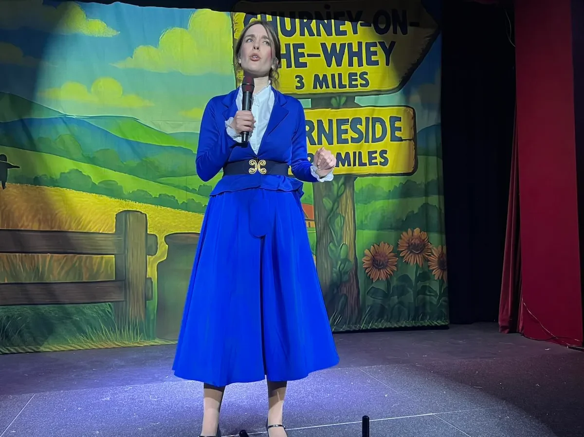 Performer and Rowena Ratzi in a blue suit holding a microphone while reporting the News