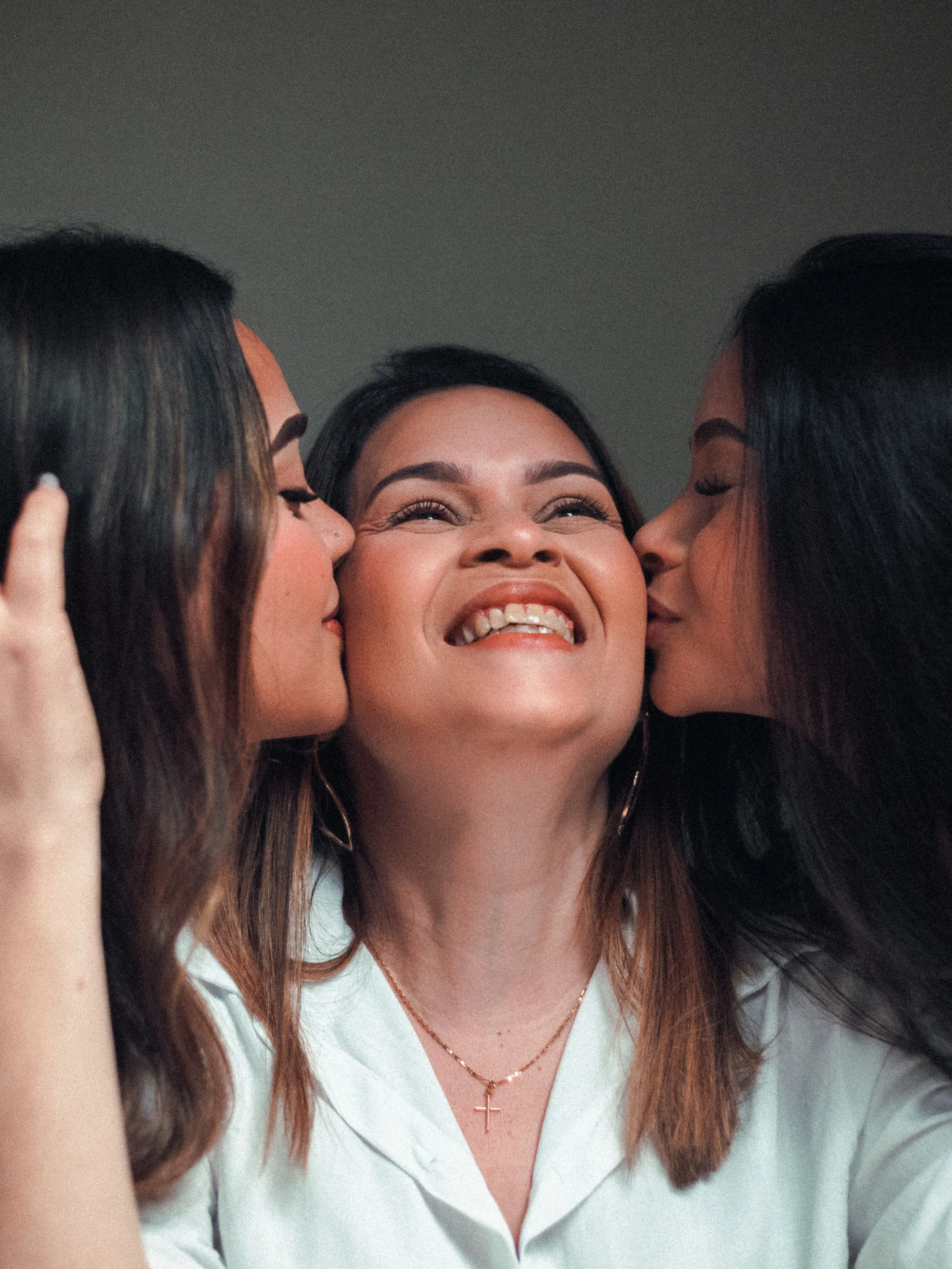 Therapy in Kitchener, A woman with dark hair smiling while being kissed on each cheek by two other women, one with dark hair and one with brown hair, all in close-up shot.