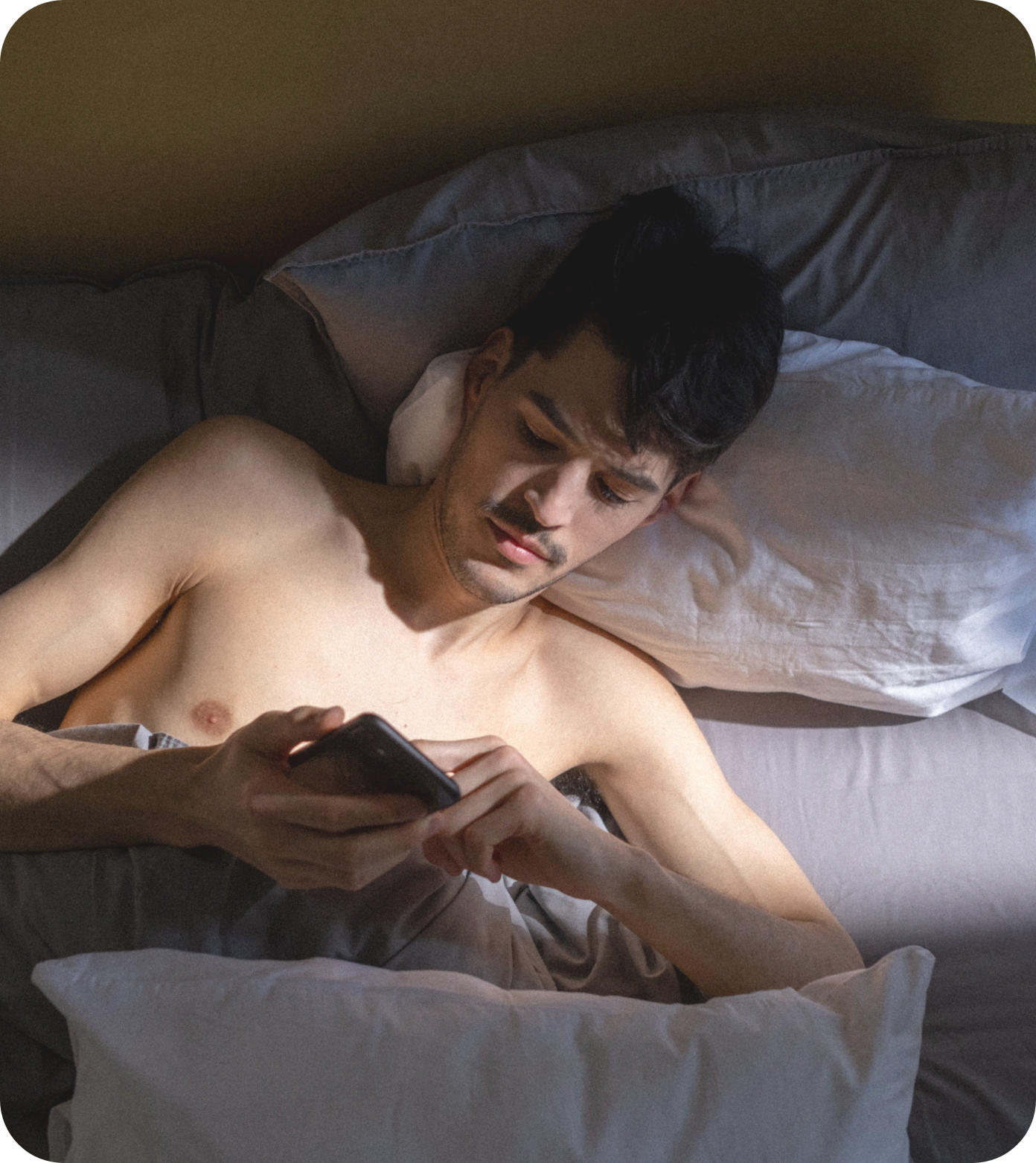 A shirtless man with dark hair lying in bed, looking at a smartphone.
