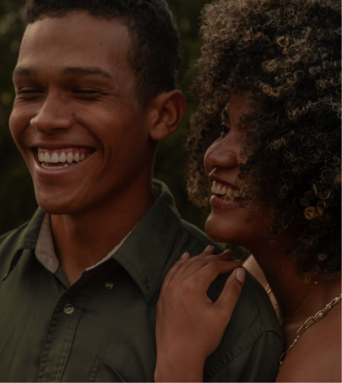 Two people smiling and enjoying each other's company, one with short dark hair and the other with curly hair, outdoors during daytime.