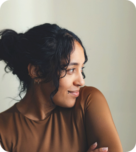 A woman with dark curly hair tied back, looking to her left with a smile, wearing a brown shirt and standing against a plain light-colored background.