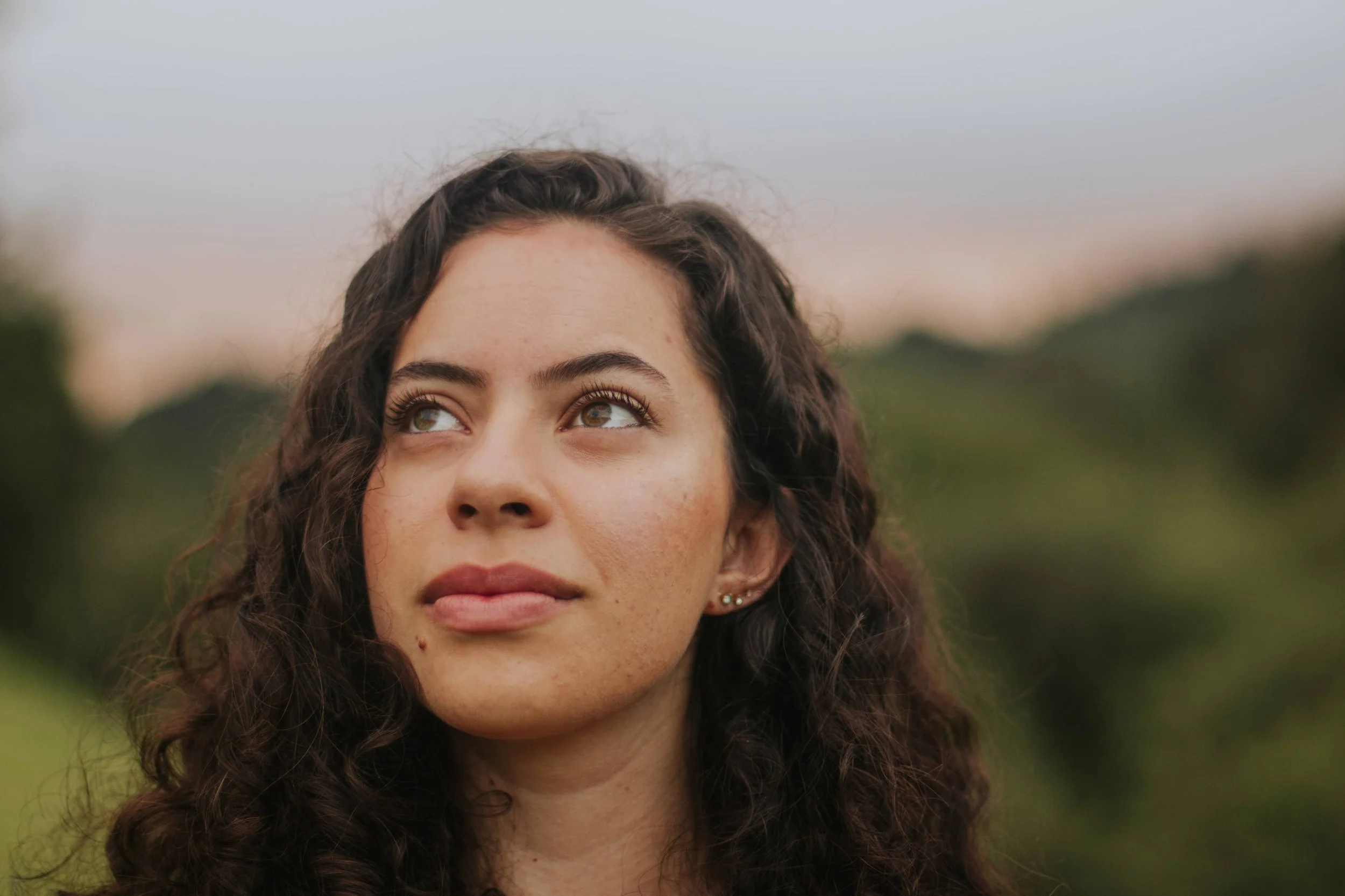 A young woman with dark curly hair looks upward with a neutral expression. She has light brown eyes, light skin, and is wearing small earrings. The background shows a blurred outdoor setting with green foliage and a soft sky.
