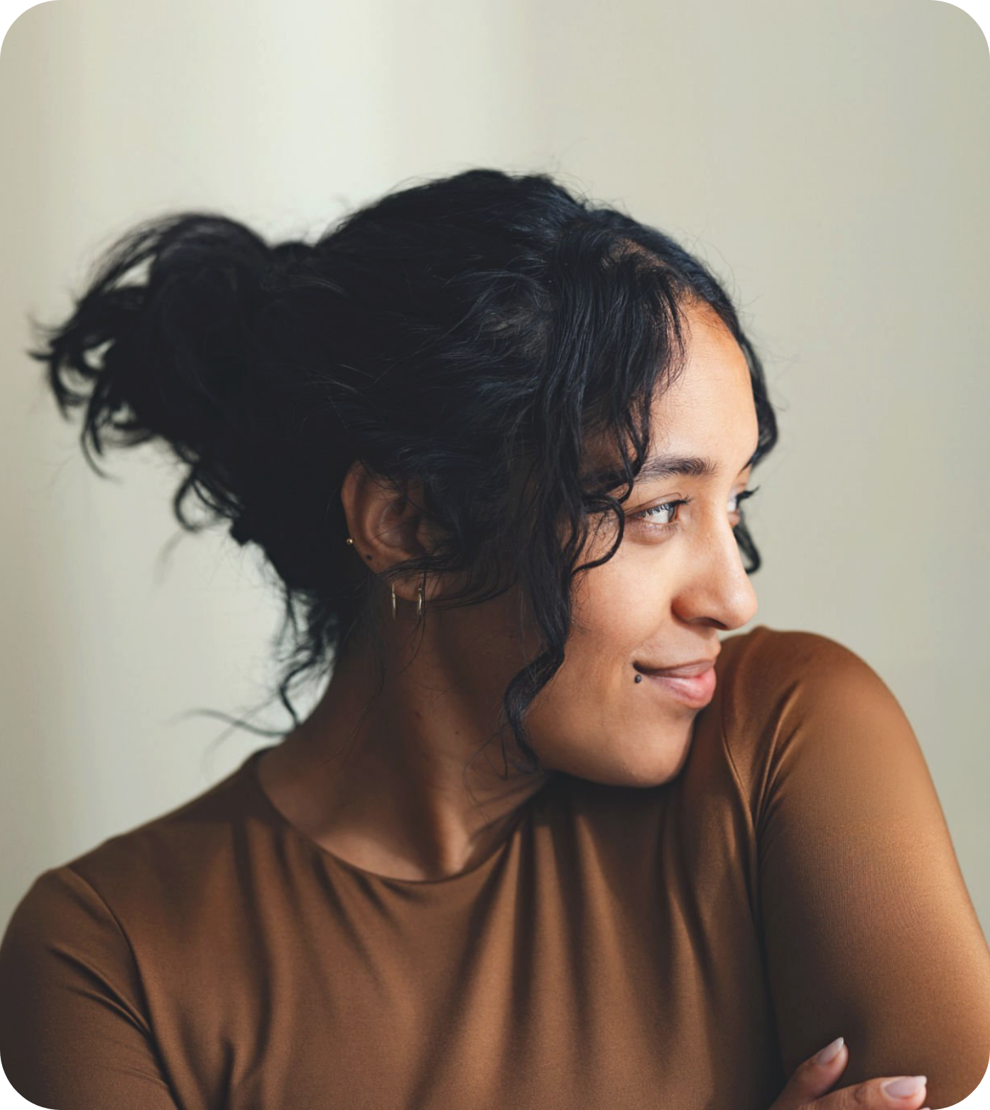 A woman with short curly black hair and a side profile, wearing a brown top and small earrings, smiling gently against a plain light-colored background.