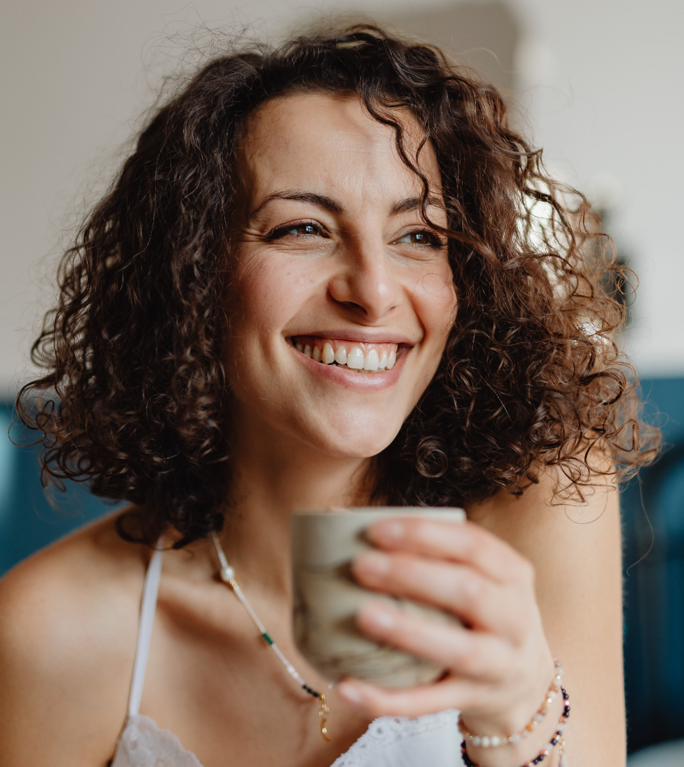 A woman with curly brown hair smiling and holding a mug.