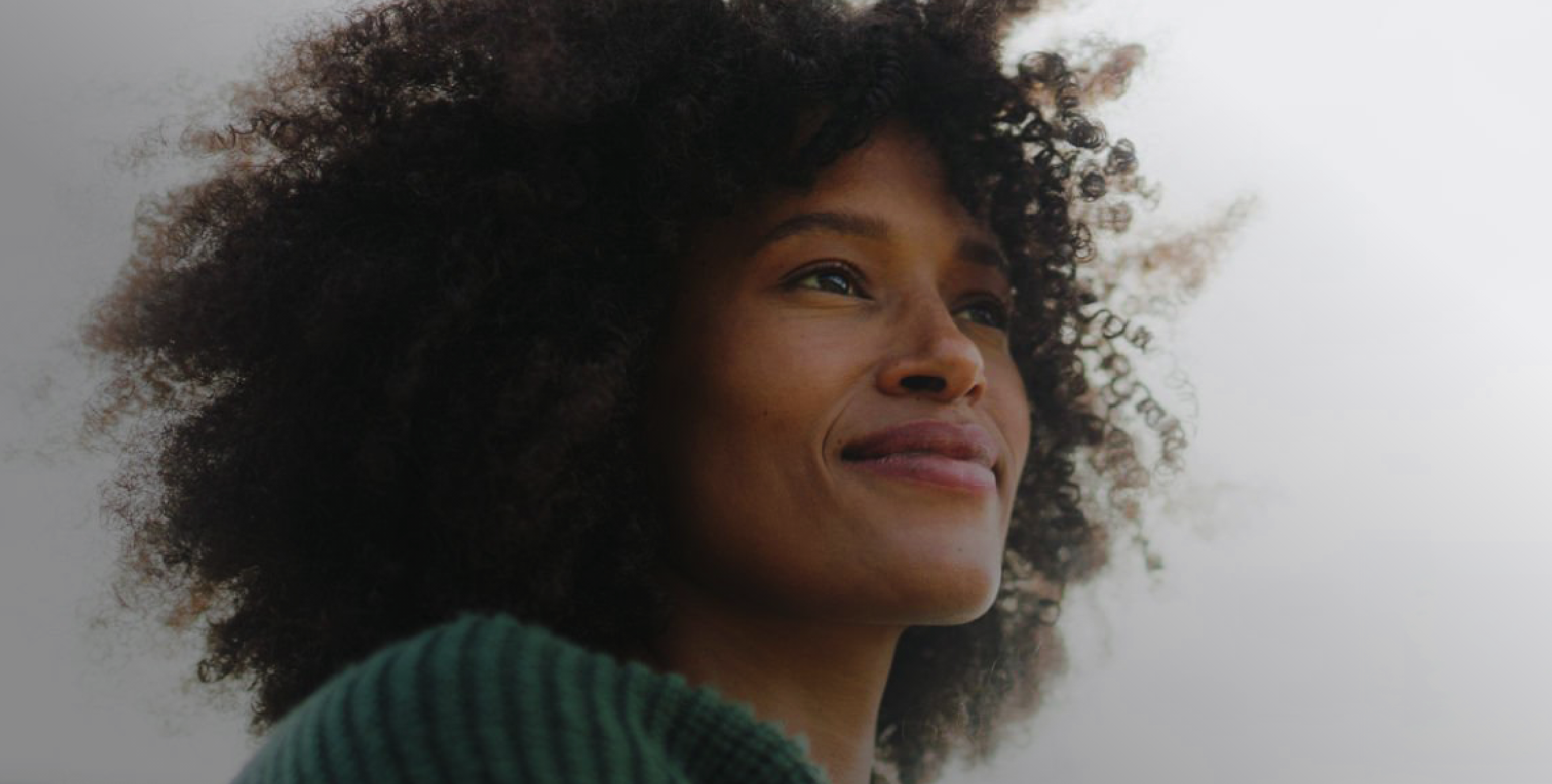 Close-up of a woman with curly hair looking to the side with a slight smile, wearing a green sweater, against a blurred, light background.