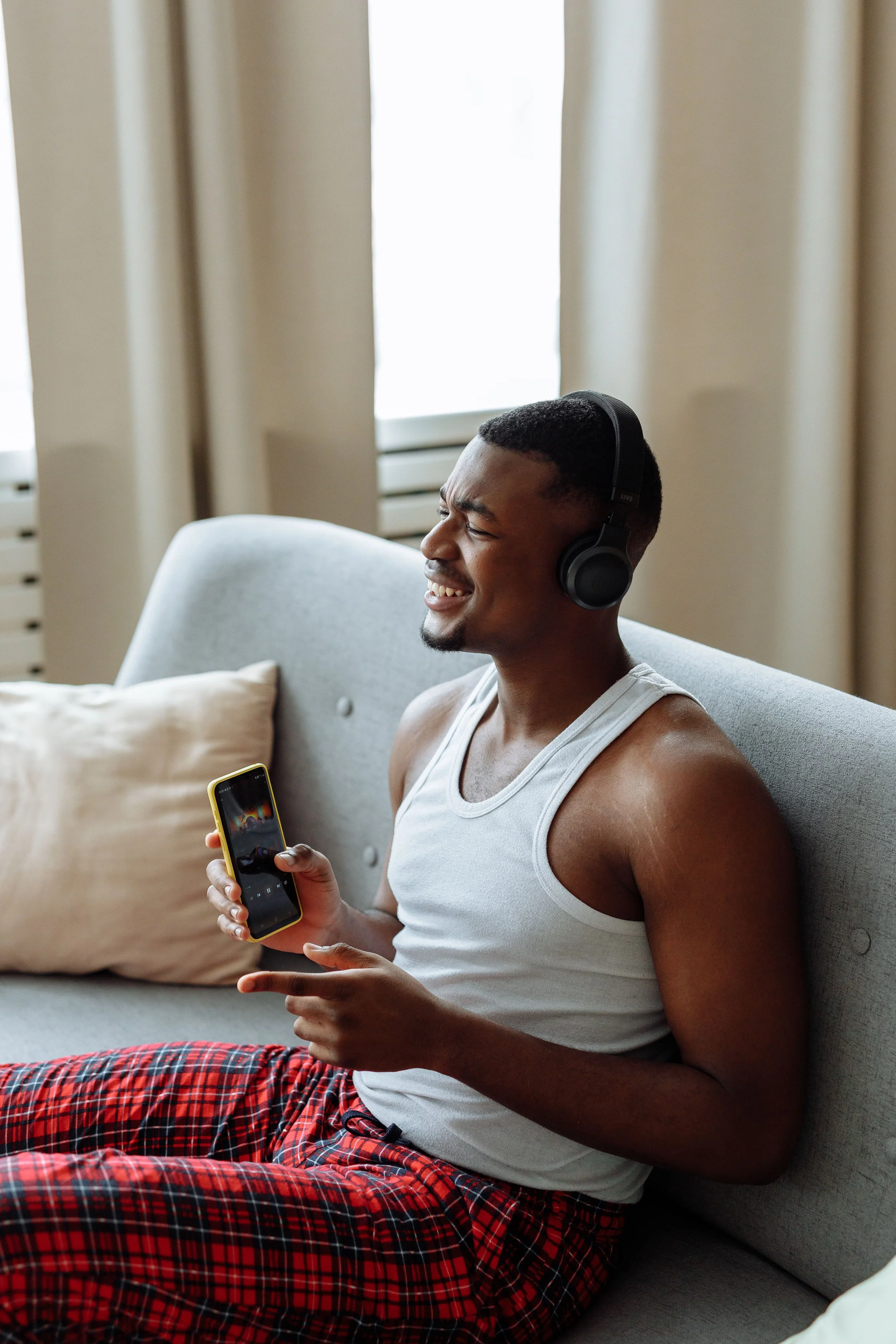 counselling Kitchener. A young man with dark hair, wearing a white tank top and red plaid pajama pants, sitting on a gray couch in a living room. He is smiling, wearing black headphones, and holding a smartphone.