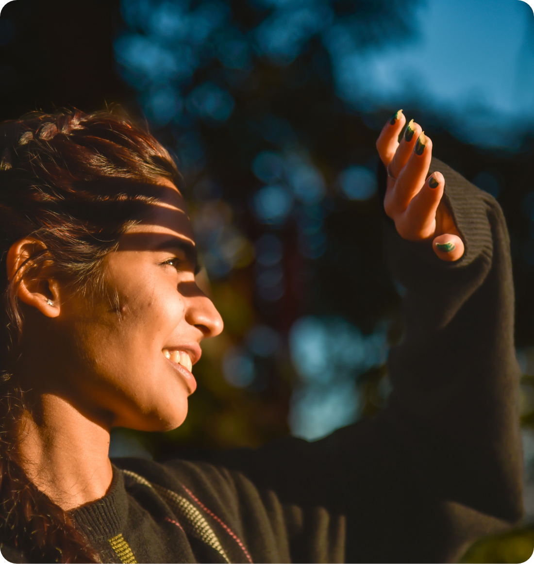 A young woman smiling and squinting in sunlight, holding her hand up with painted nails in front of her face, outdoors during evening or dusk.